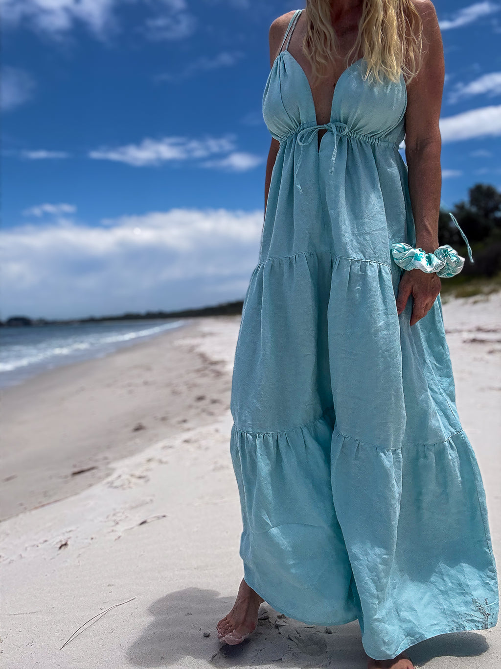 Woman in a light blue dress standing on a beach with a blue sky and ocean in the background wearing a large blue silk scrunchie on her wrist.