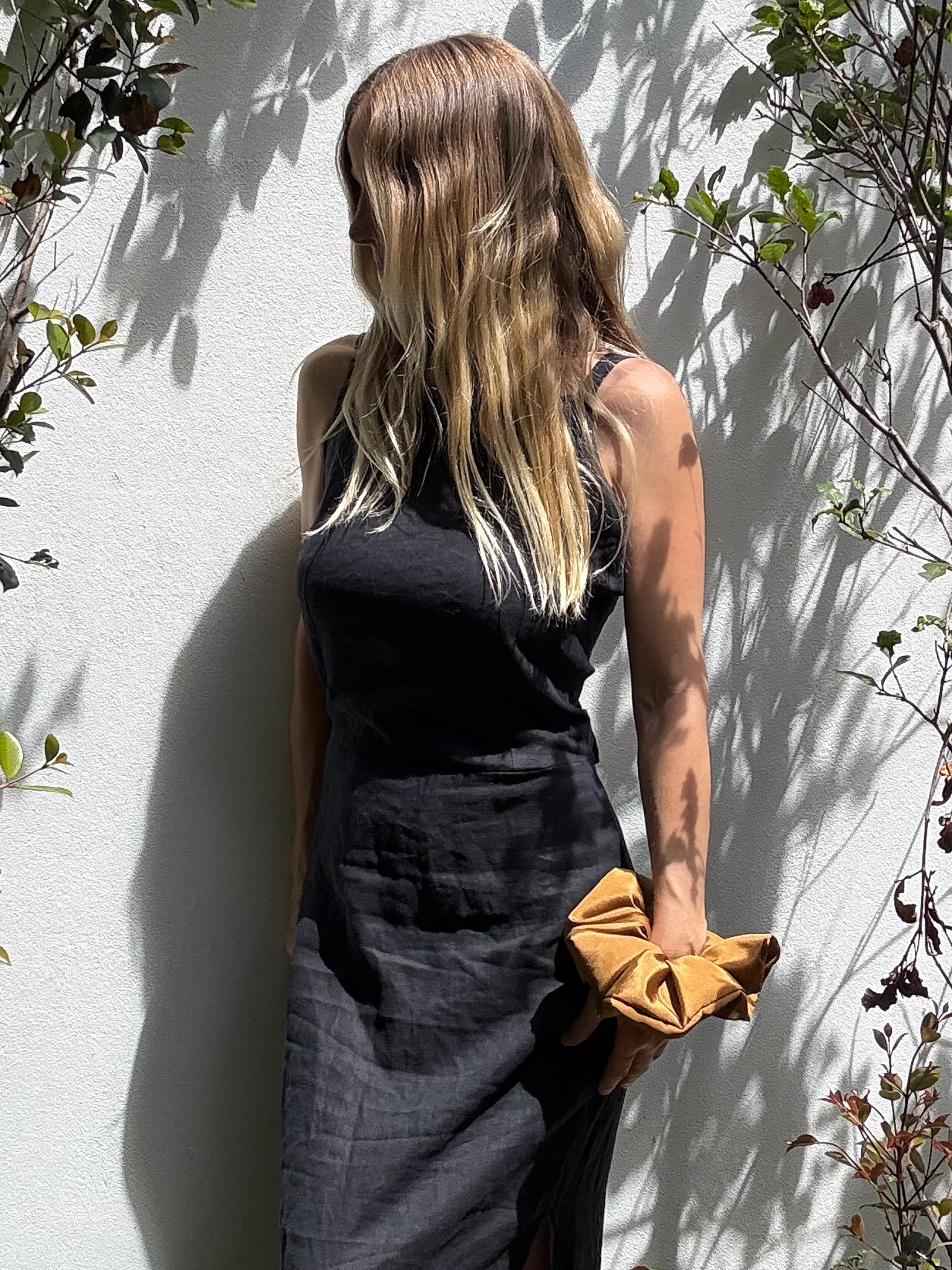Person wearing a black sleeveless dress and a large bronze coloured scrunchie on her wrist against a light wall with plants.