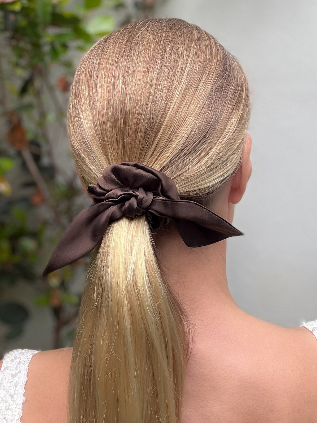 Person with blonde hair tied into a ponytail with a brown scrunchie against a blurred natural background