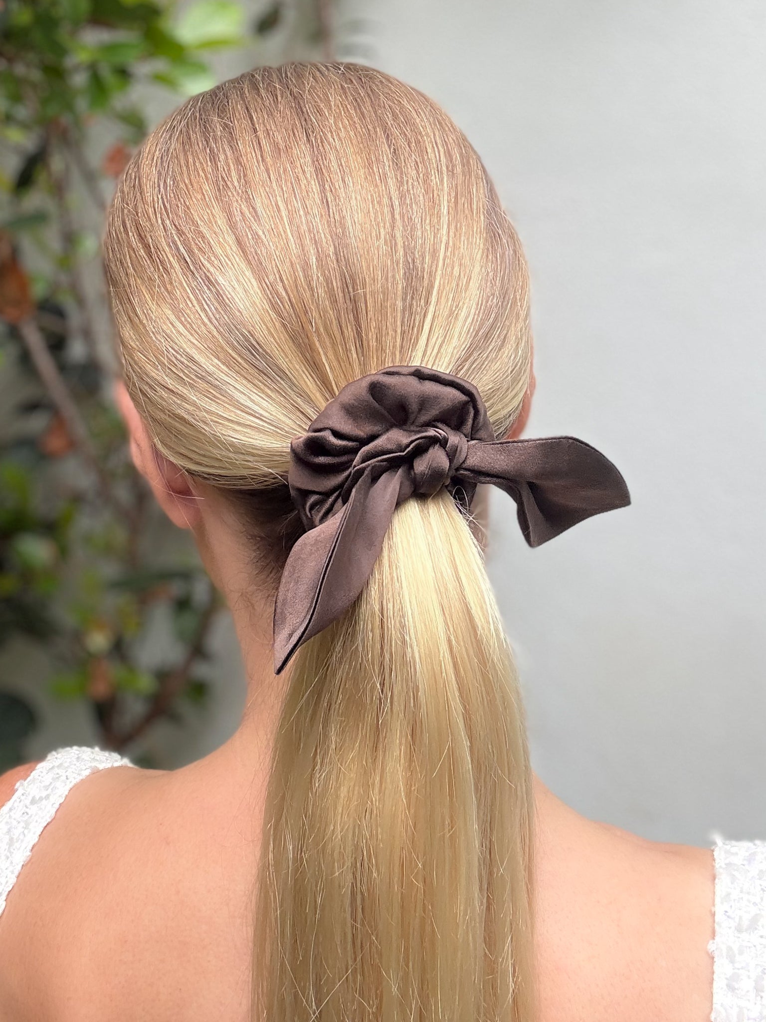 Blonde hair tied into a ponytail with a brown scrunchie against a neutral background