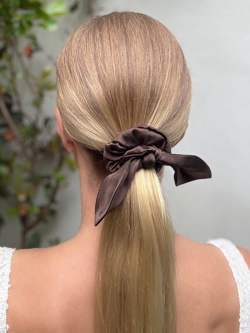 Person with long blonde hair tied in a ponytail with a brown scrunchie, against a neutral background.