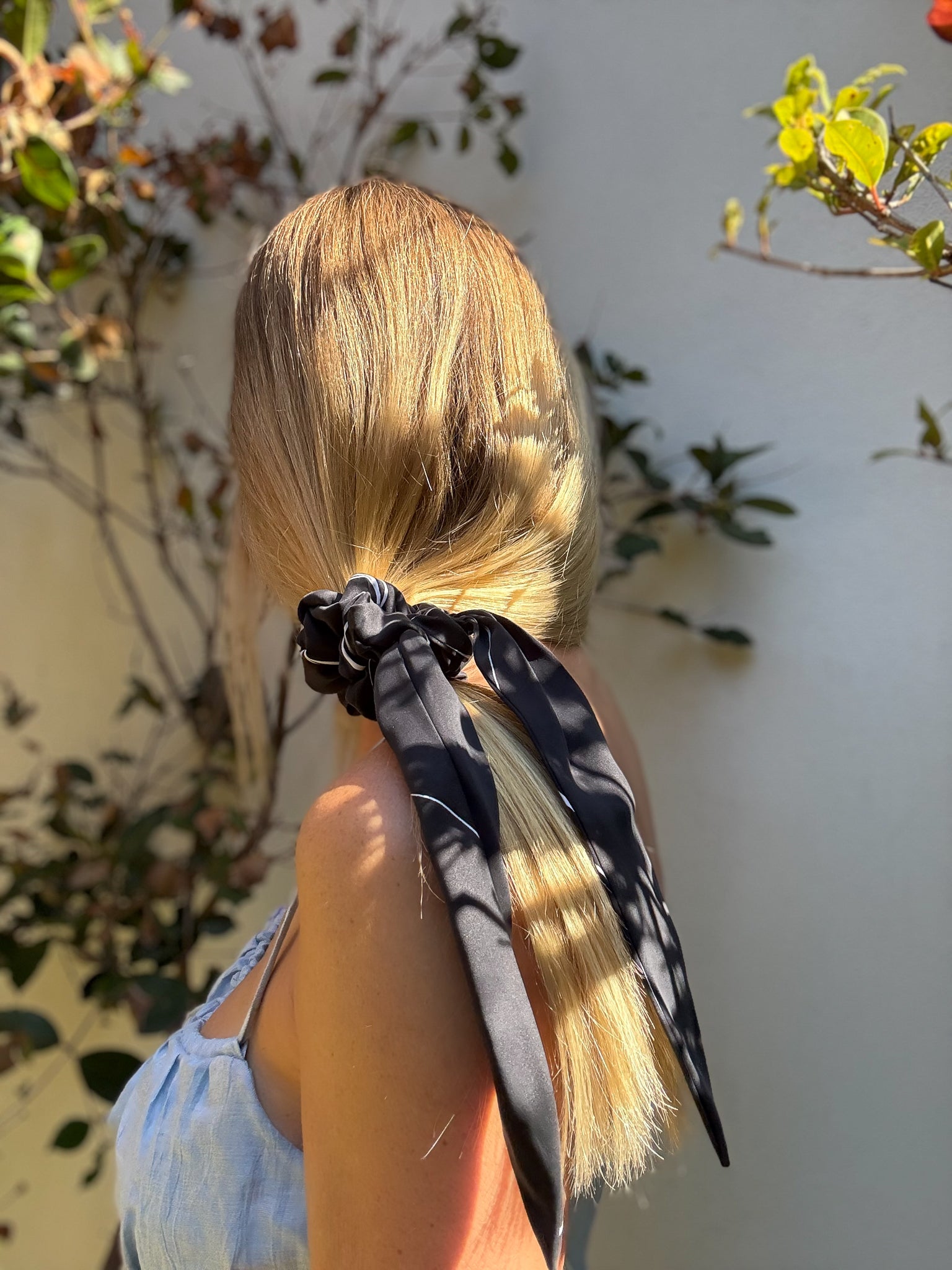 Person with blonde hair tied back with a black scrunchie featuring long tails, against a natural background.