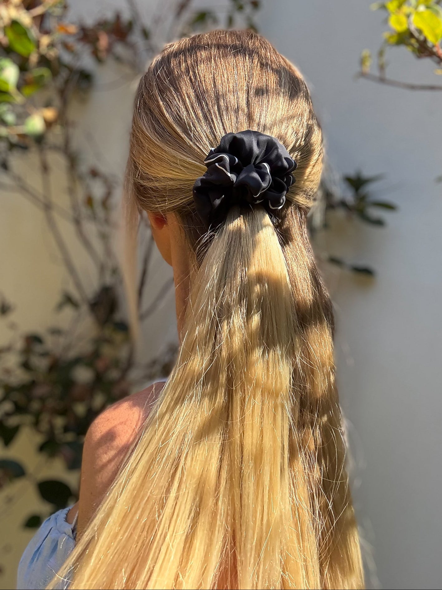 Person with long blonde hair tied back with a black silk scrunchie, outdoor setting.