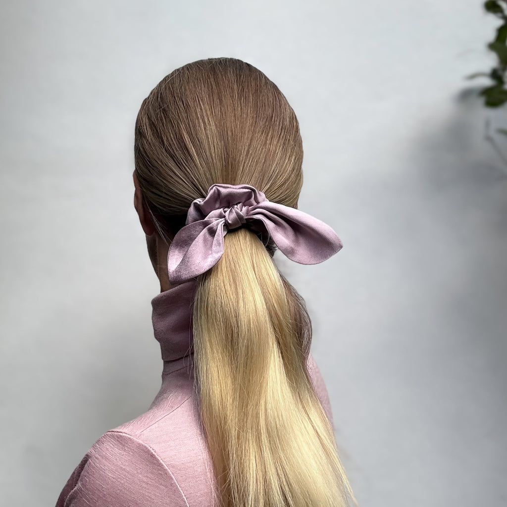 Person with long blonde hair tied in a ponytail with a purple silk scrunchie against a light background