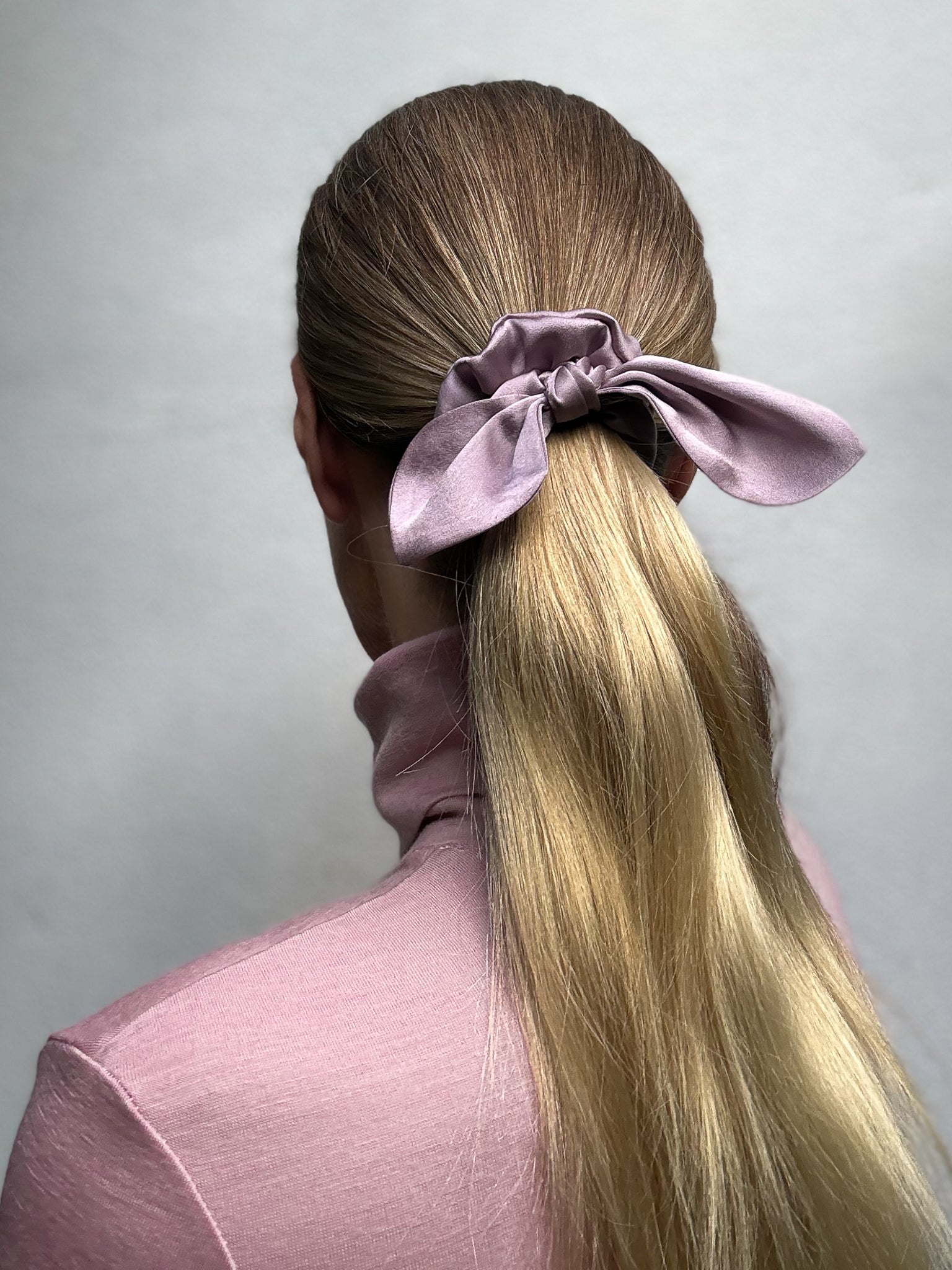 Person with long blonde hair tied in a ponytail with a purple scrunchie against a light gray background