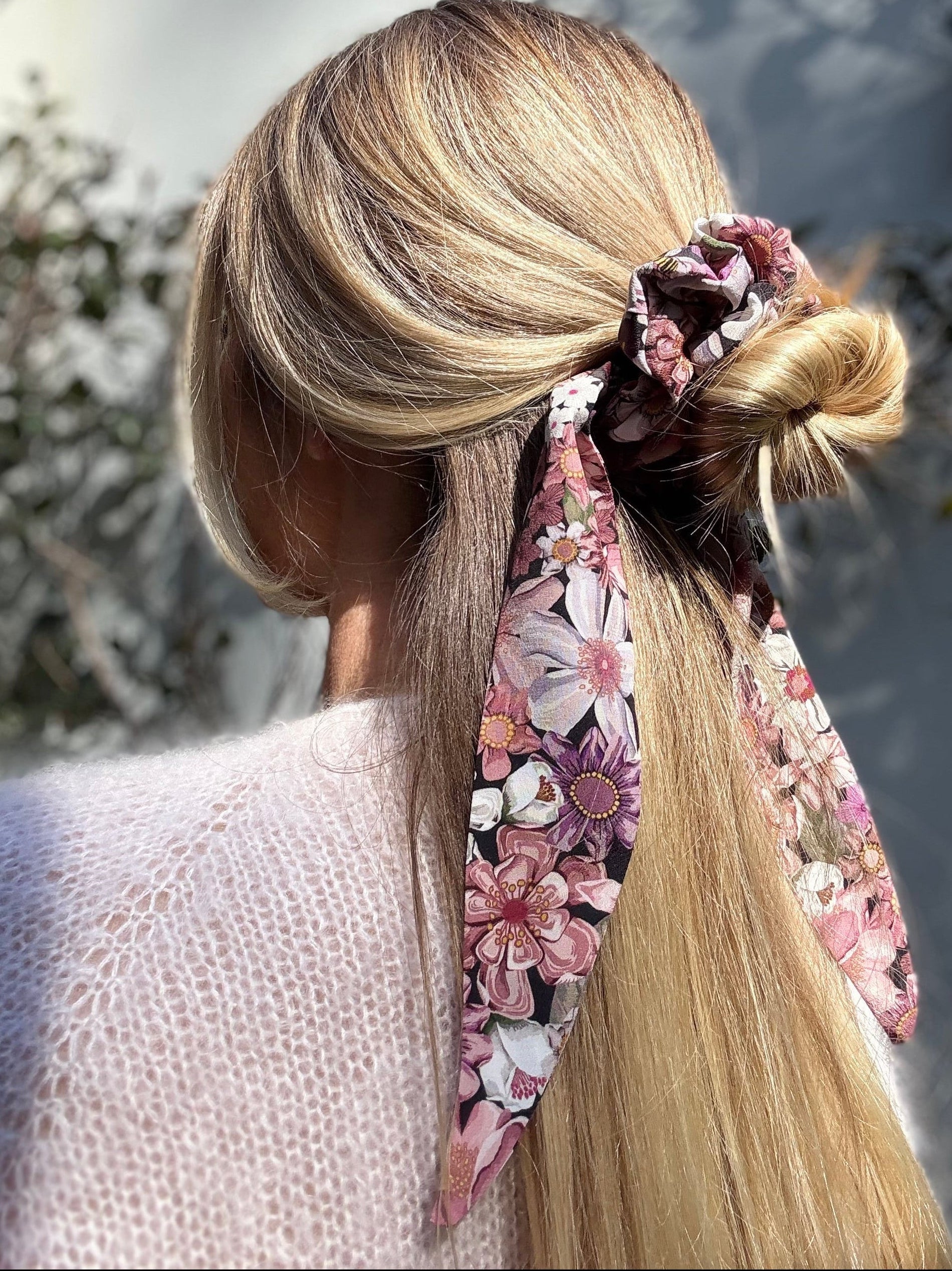 Woman with blonde hair tied back using a floral silk scrunchie with a blurred natural background