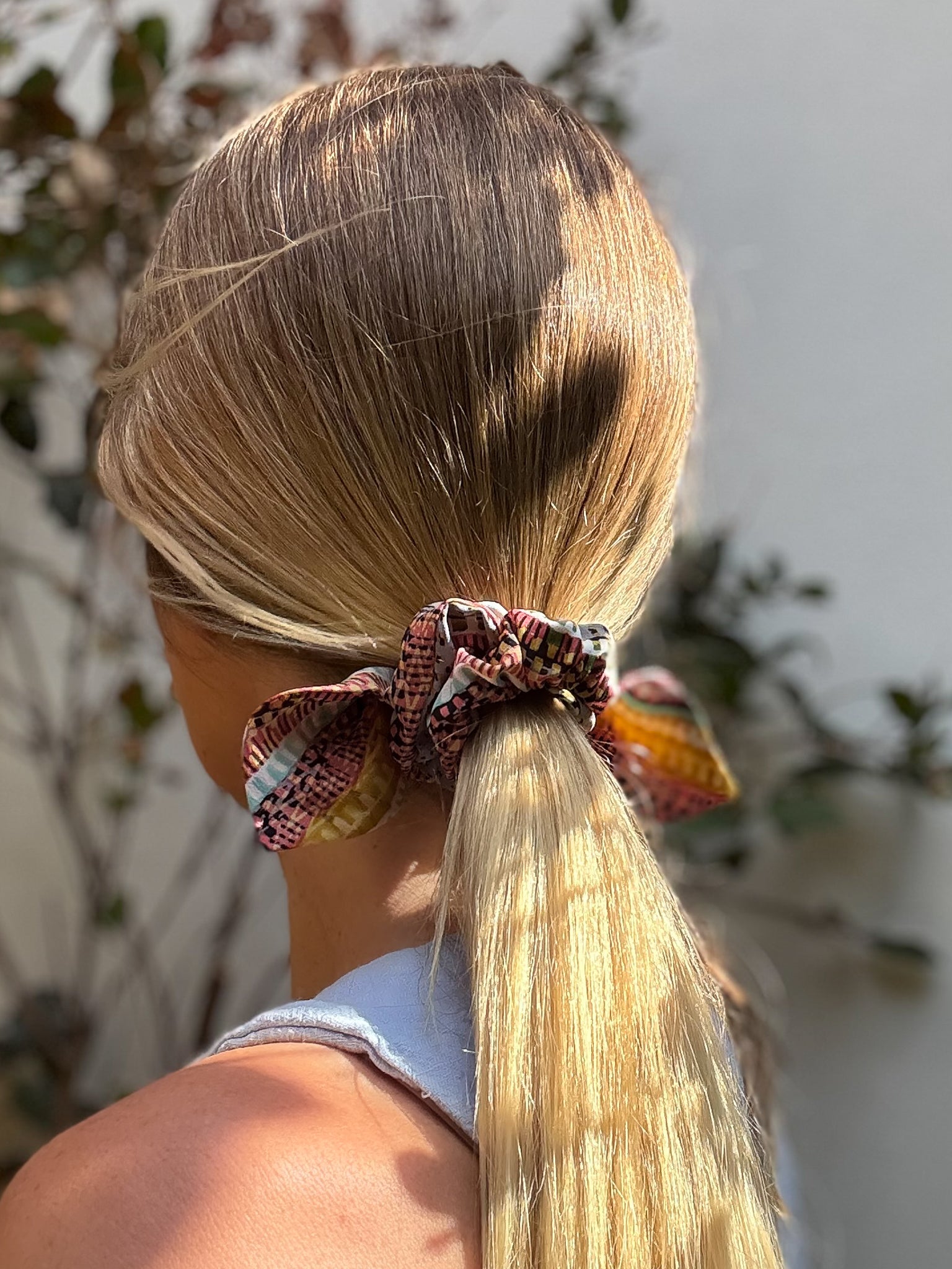 Person with blonde hair tied back with a small silk scrunchie, blurred outdoor background