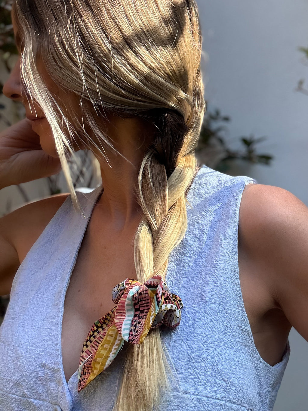 Person with braided hair wearing a colorful silk scrunchie with detachable bow, outdoors.