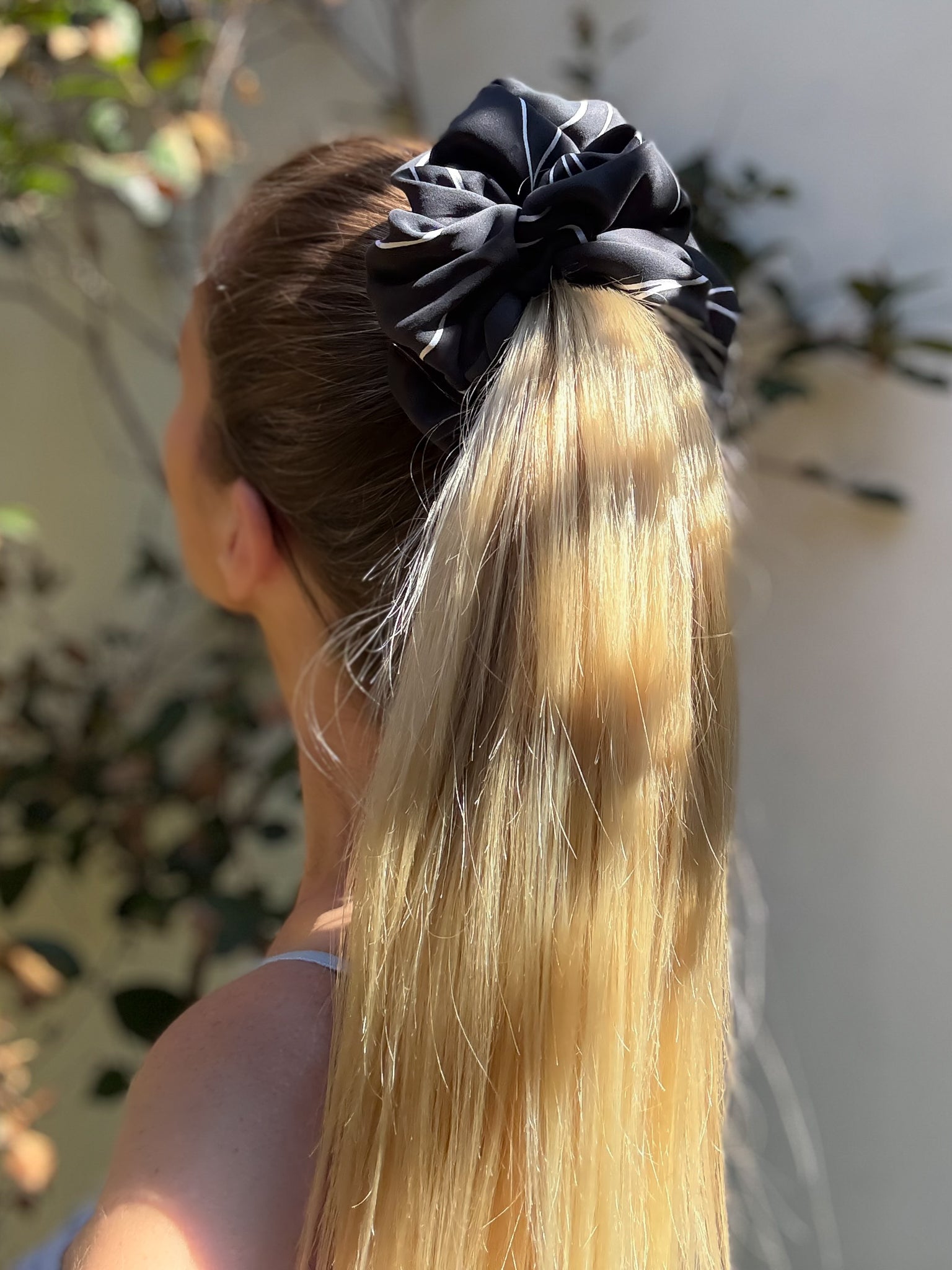 Person with blonde hair tied in a ponytail with a black scrunchie against a blurred outdoor background
