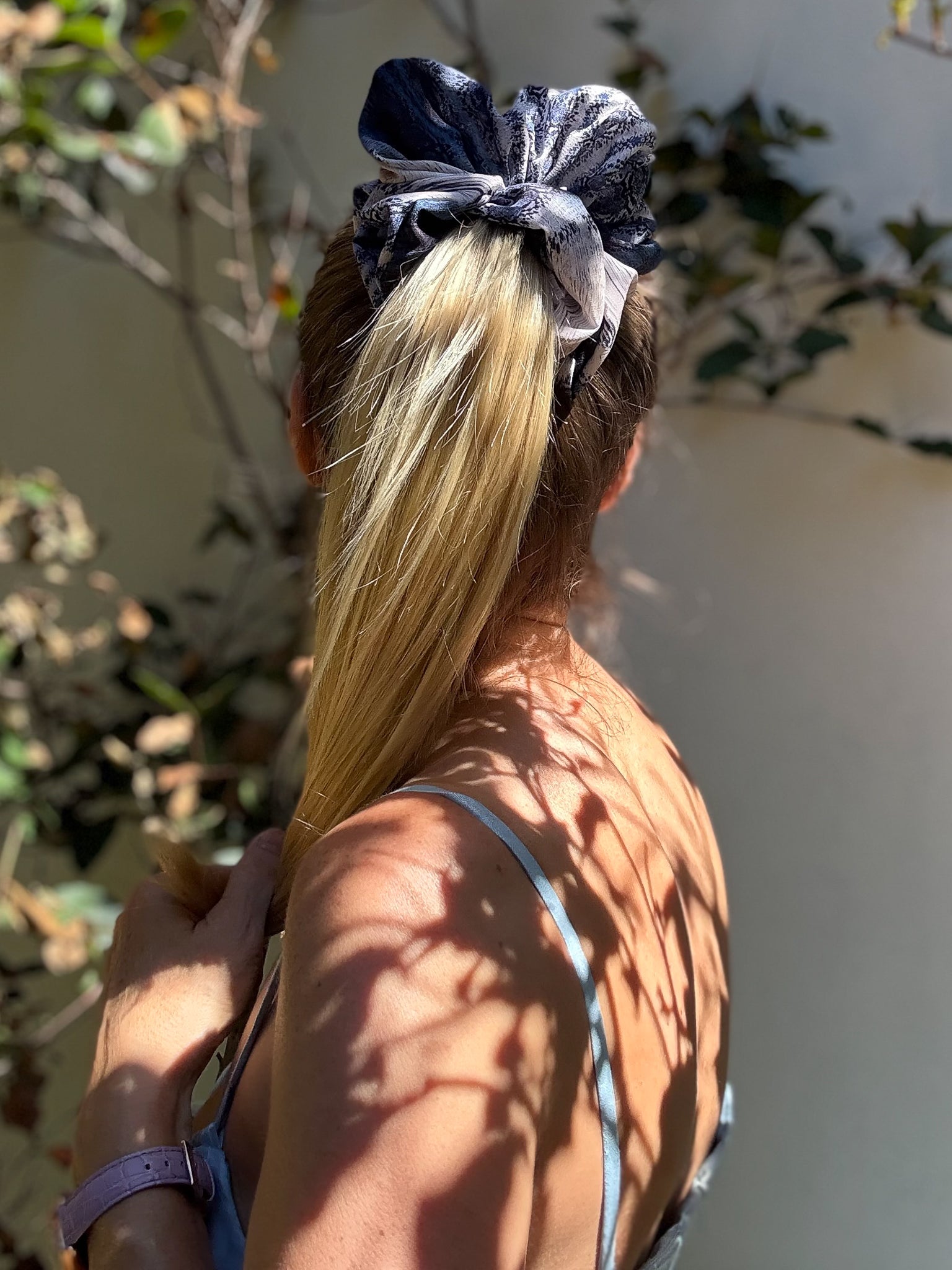 Person with a blue scrunchie in their hair, standing outdoors with plants in the background