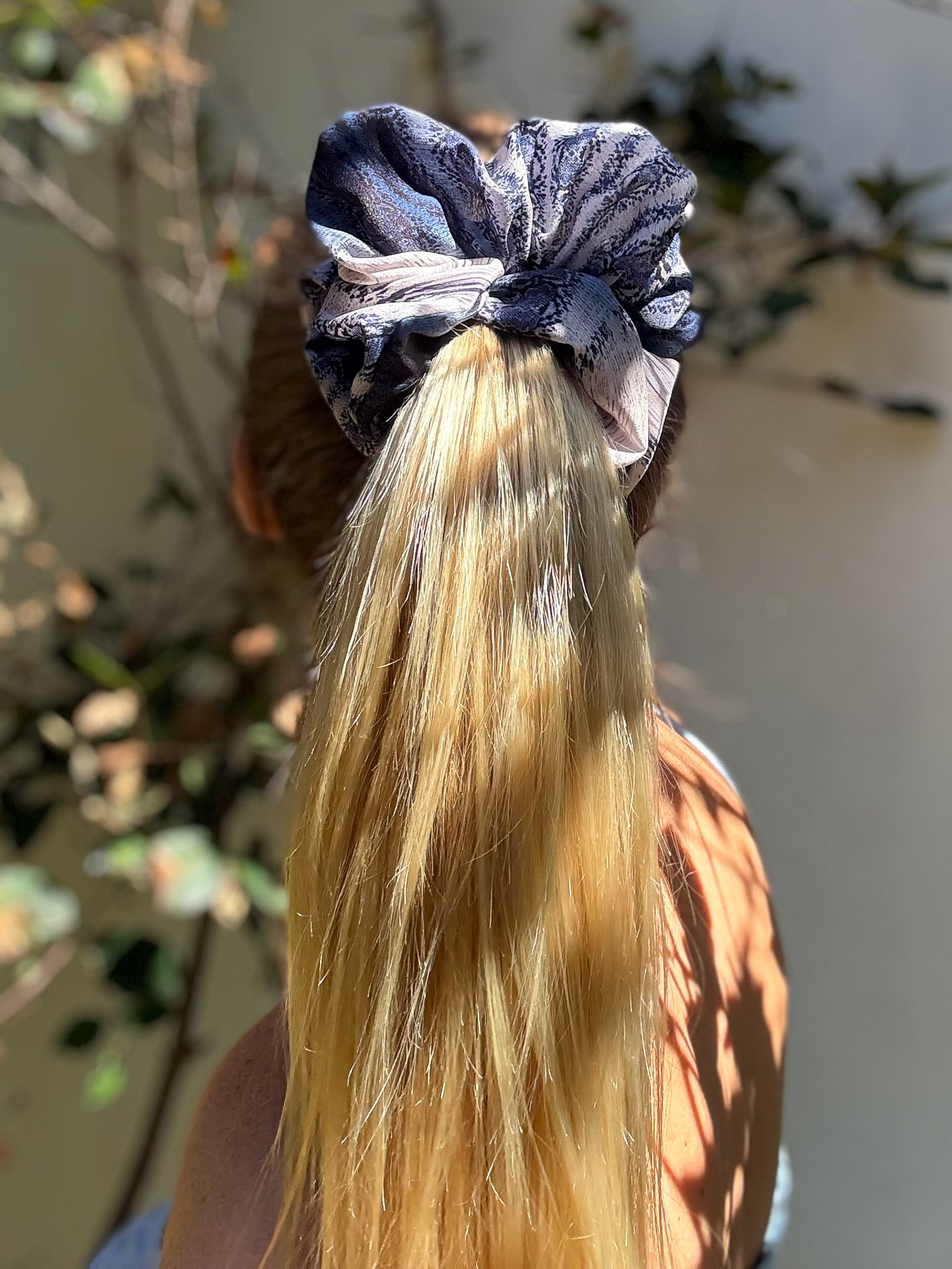 Person with long blonde hair tied up with a blue scrunchie against a blurred outdoor background