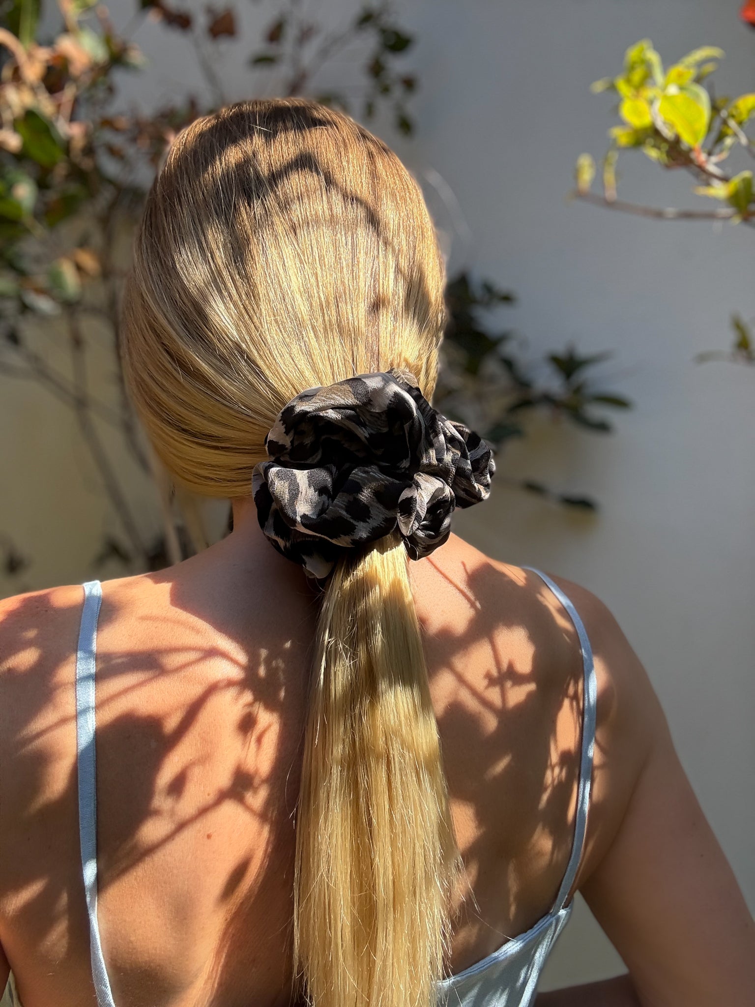 Person with blonde hair tied in a ponytail with a animal print scrunchie, standing outdoors with plants in the background.