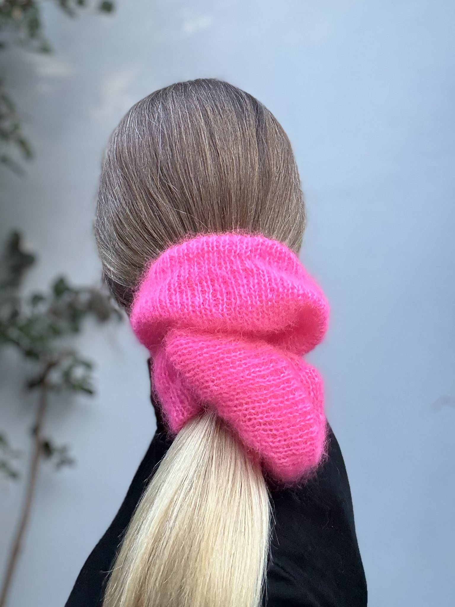 A bright pink fluffy knit ponytail hair tie worn around a blonde woman's ponytail, shown from behind.
