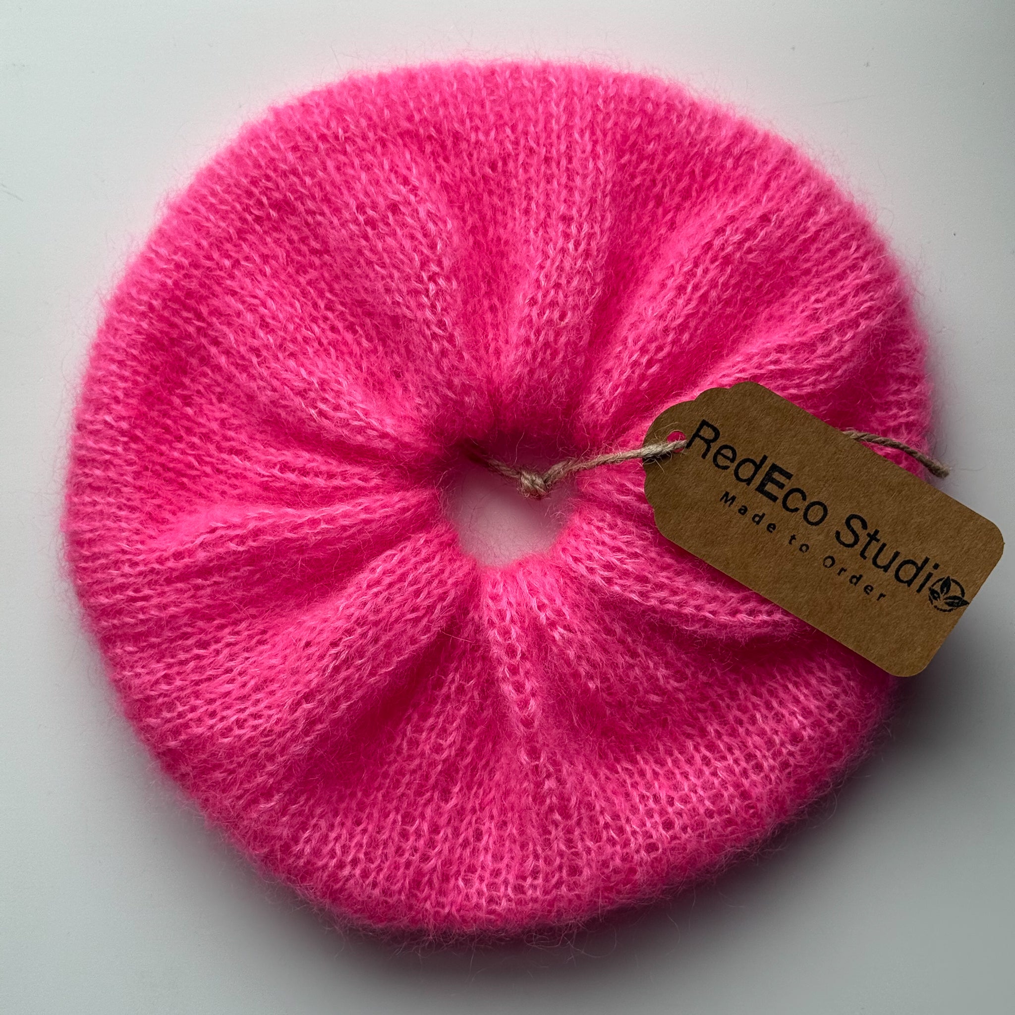 A bright pink knitted hair scrunchie with a brown paper hangtag that reads 'RedEco Studio Made to Order', shown on a plain light background.
