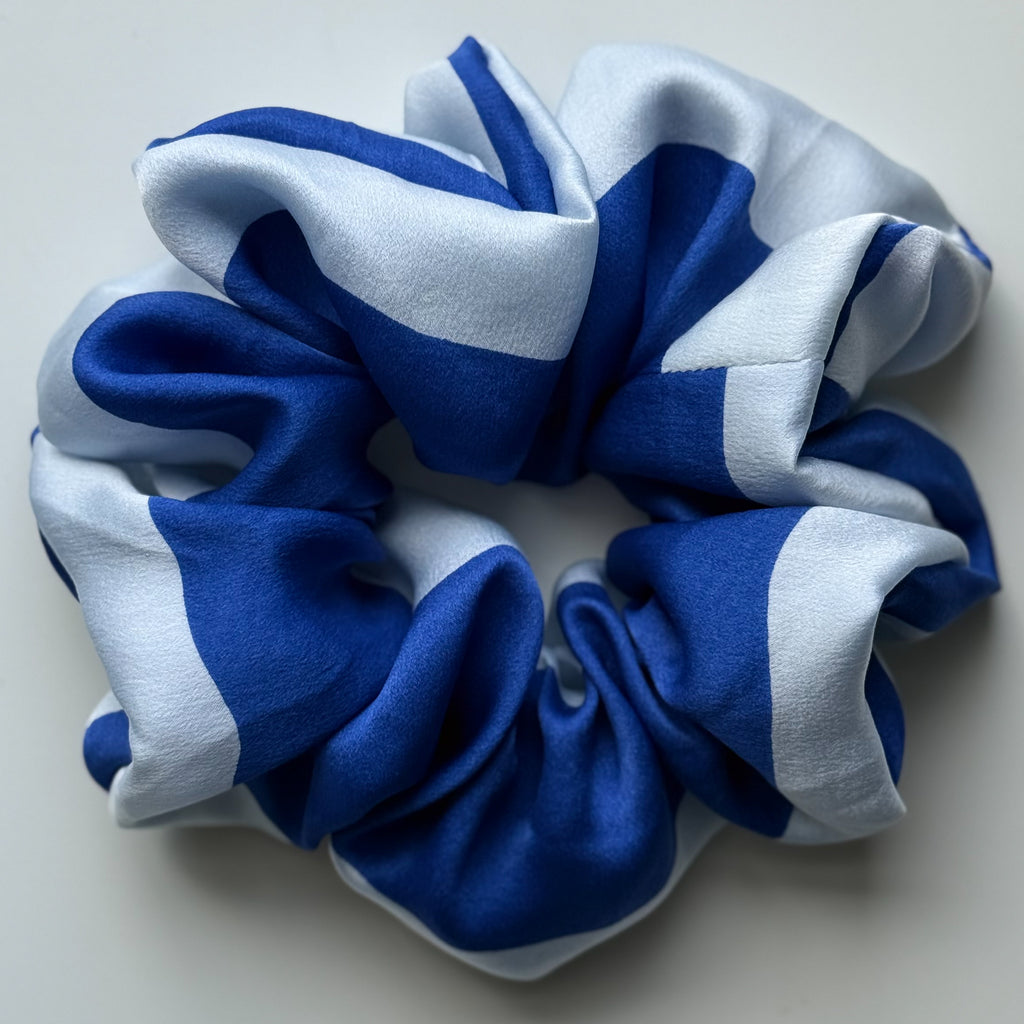 A silk hair scrunchie with a bold blue and light blue striped pattern, photographed on a neutral light gray background.