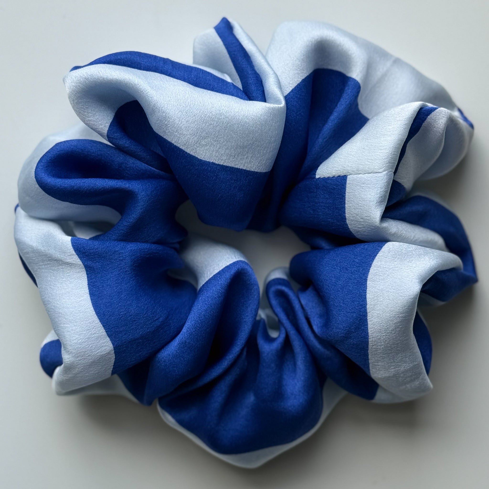A silk hair scrunchie with a bold blue and light blue striped pattern, photographed on a neutral light gray background.