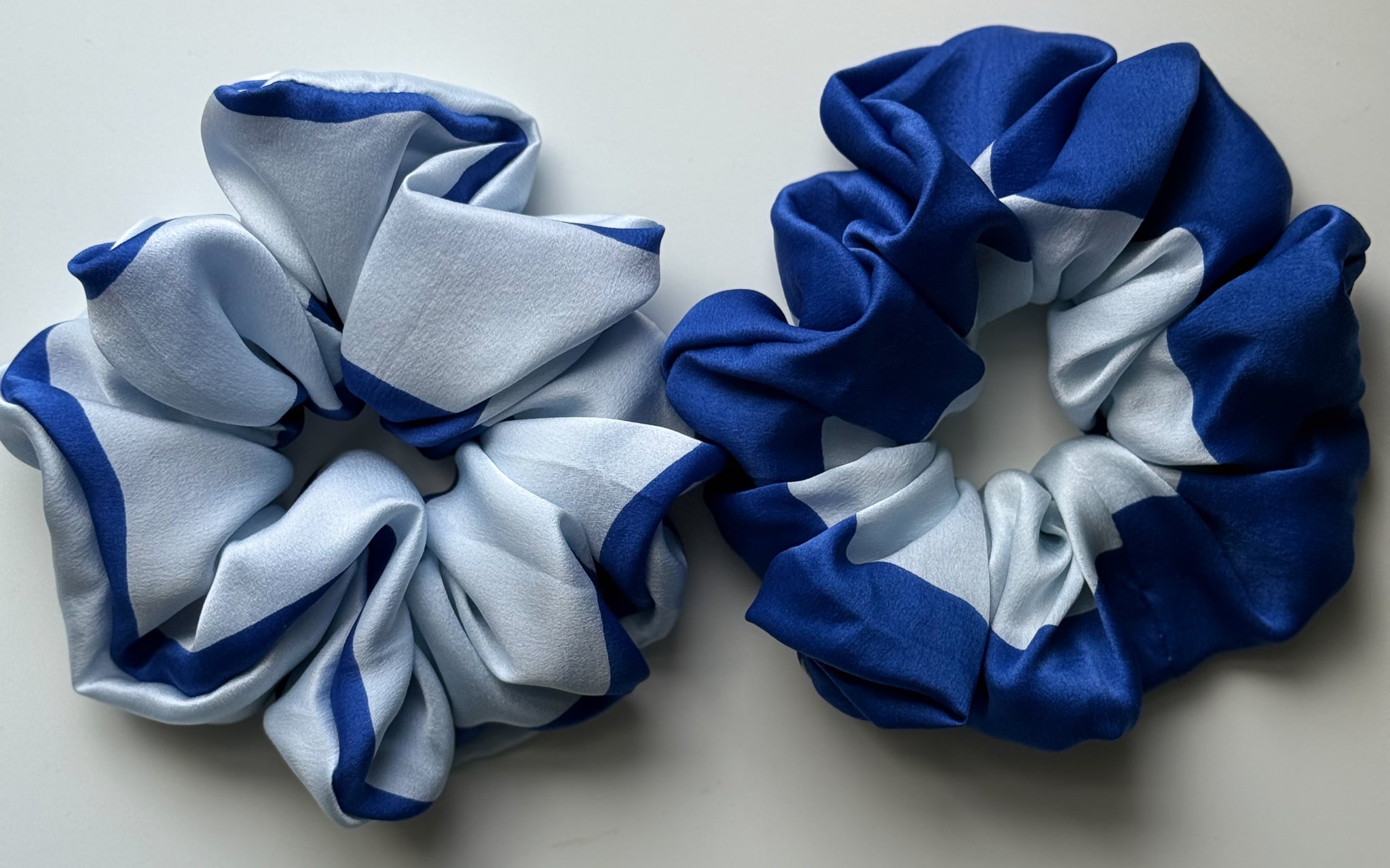 Two silk hair scrunchies on a light gray background, one in light blue and white with dark blue trim, and the other in royal blue and white with dark blue trim.