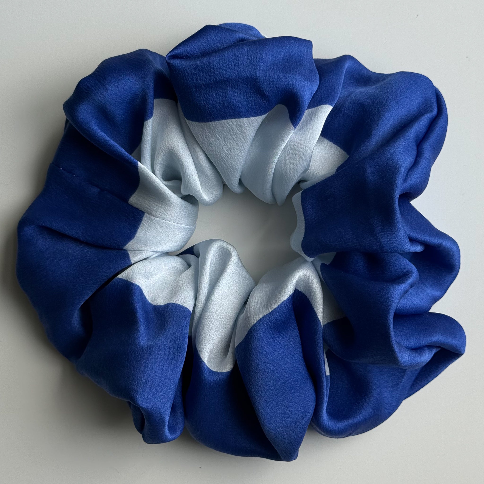 A silk hair scrunchie with a blue and white colour-blocked pattern, photographed from a top-down view on a neutral light gray background.
