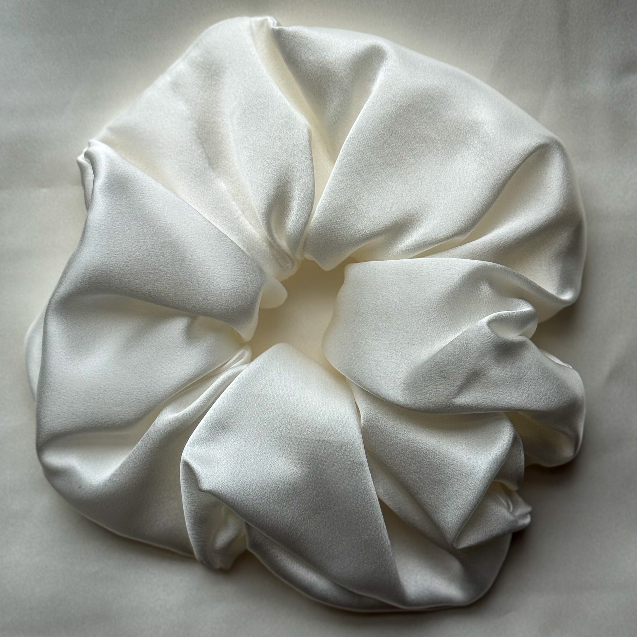 A large, round white satin hair scrunchie with soft, draped folds, photographed on a neutral light beige fabric background.