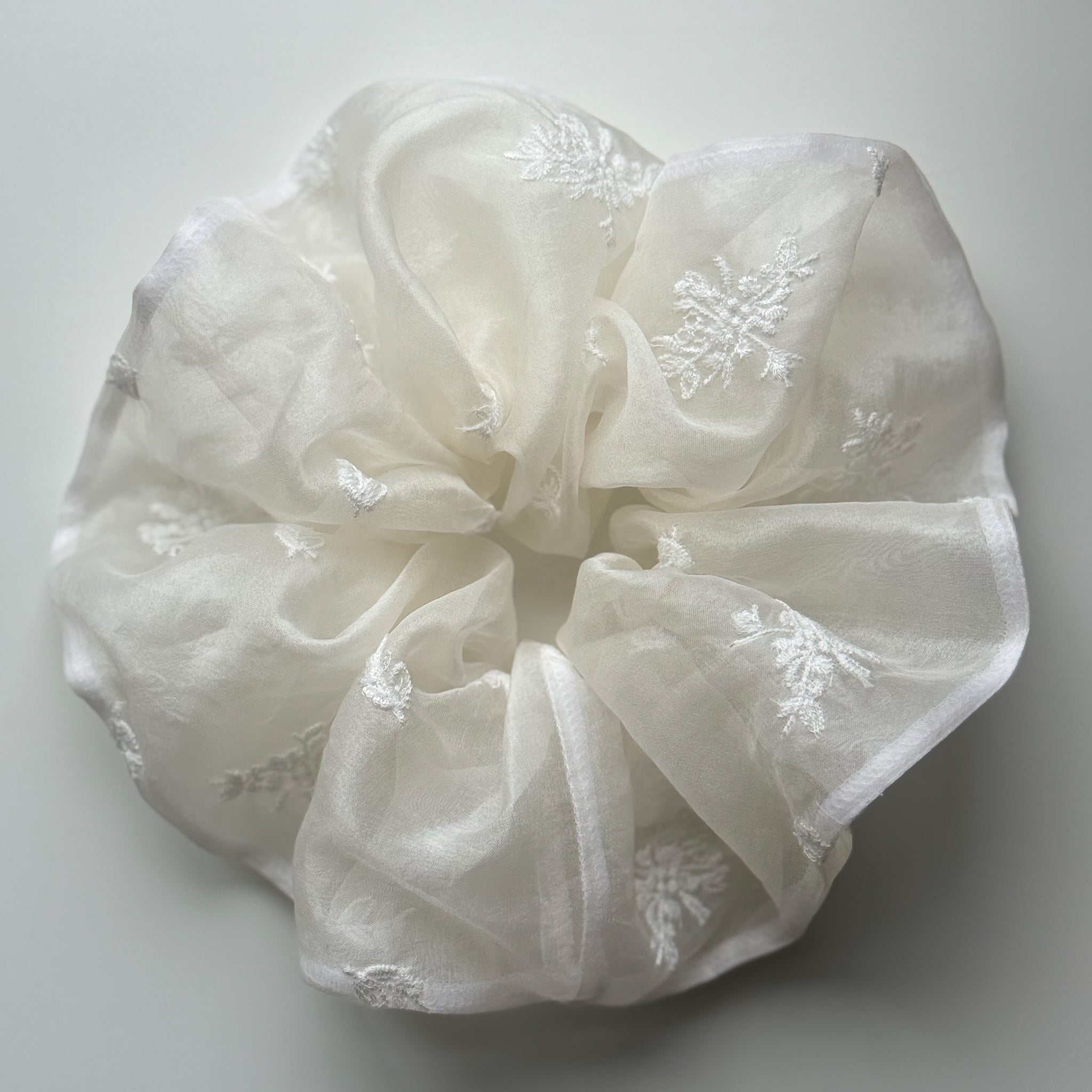 An off-white sheer organza hair scrunchie with delicate embroidered floral details, photographed on a neutral light gray background.
