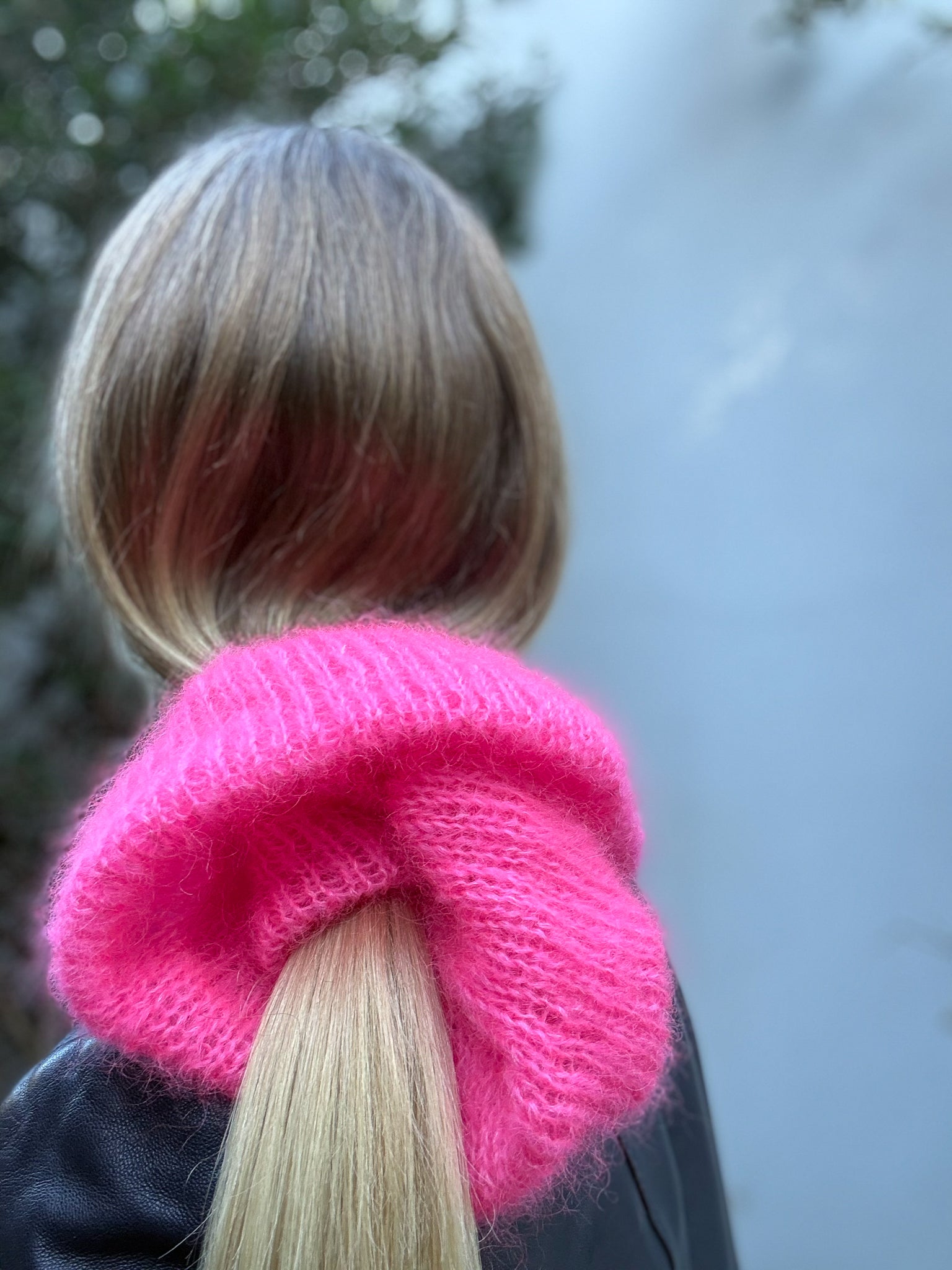 A close-up back view of a person with blonde hair in a ponytail, wearing a bright hot pink fuzzy knit scrunchie.
