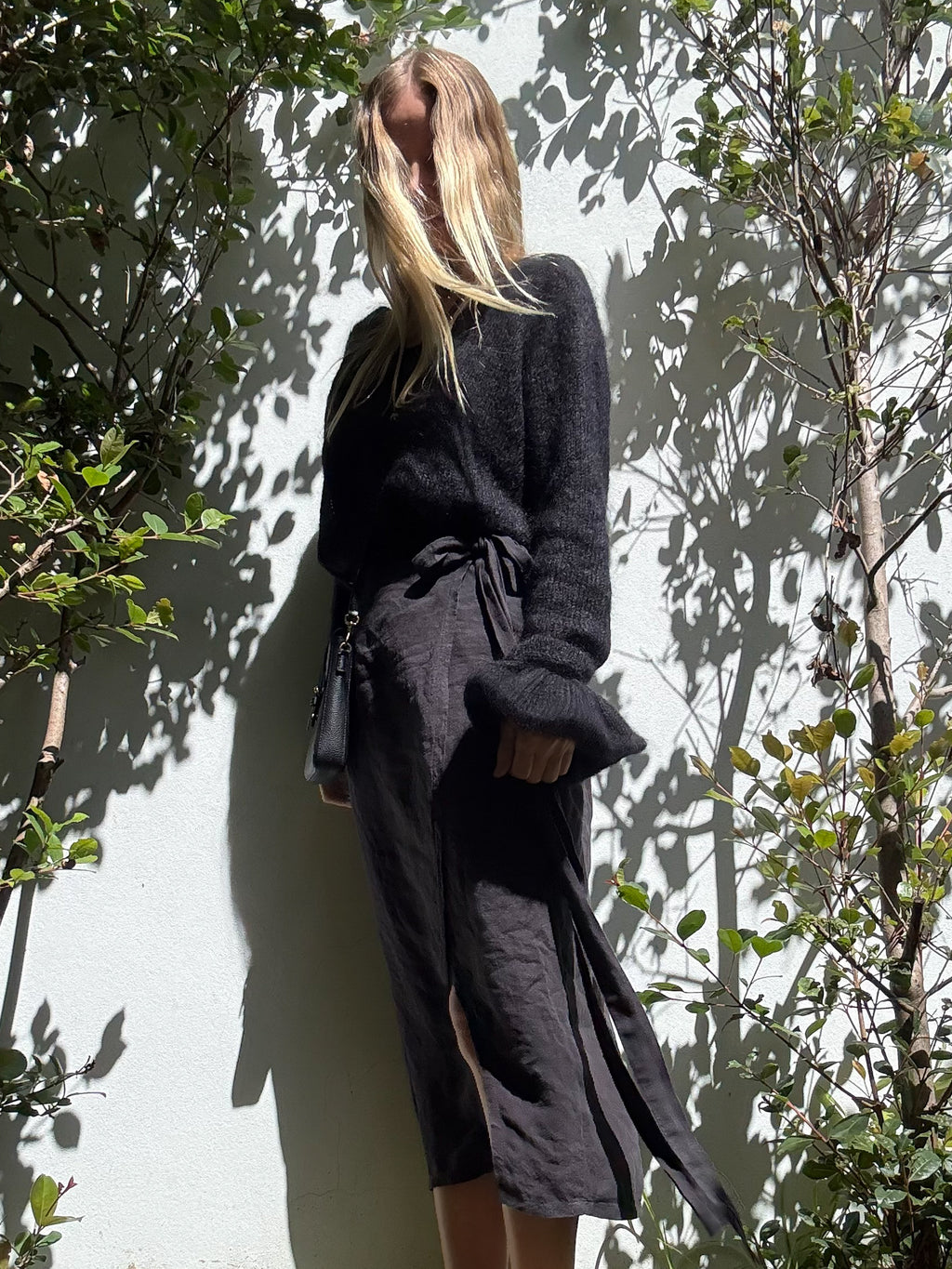 A woman stands outdoors against a white wall, wearing a black fuzzy long-sleeve top and a dark wrap-style midi skirt with a tie waist together with a matching extra large black hand-knitted scrunchie on her wrist.