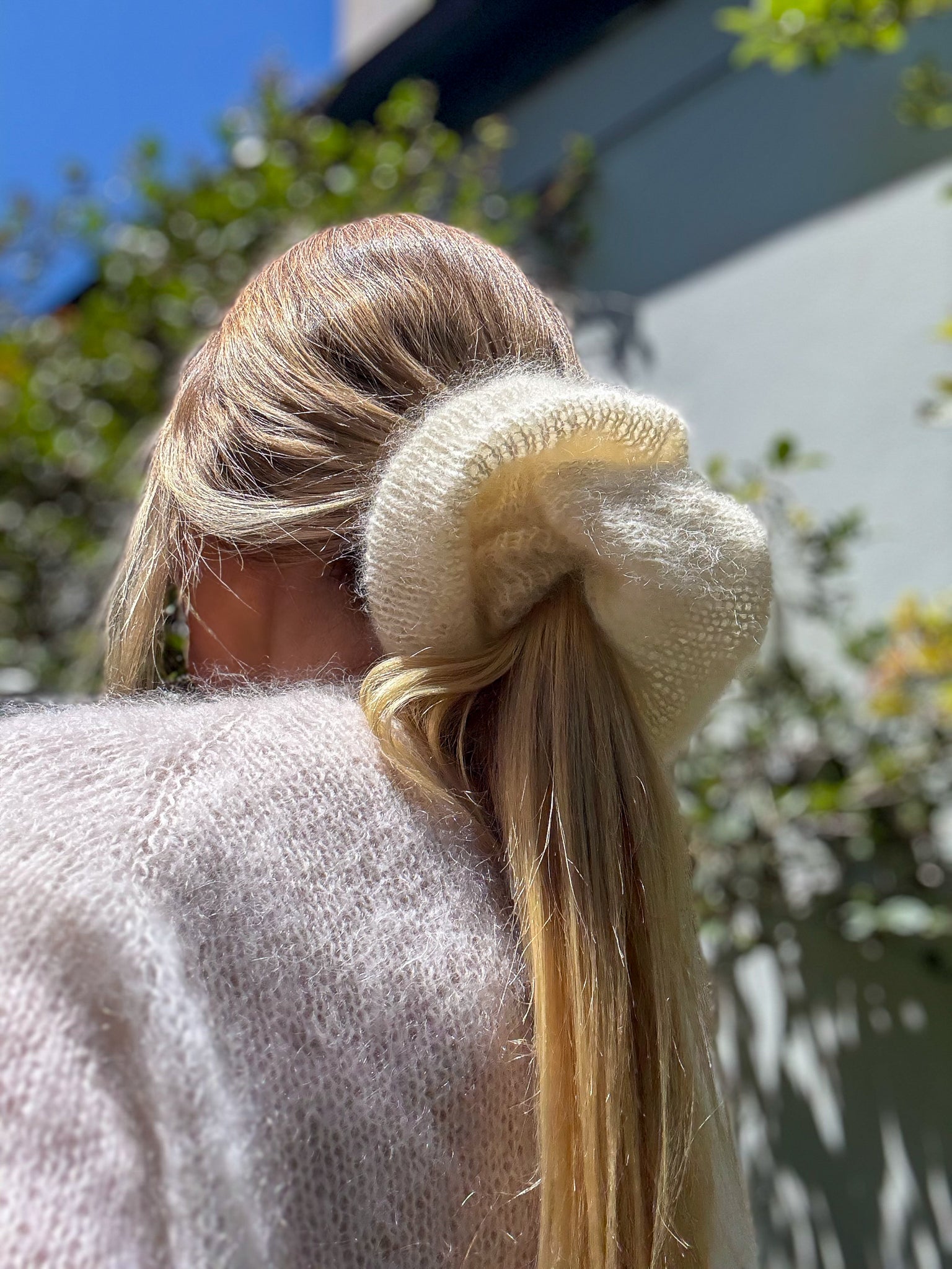 Person with a large cream hand knitted scrunchie in their hair, outdoors.