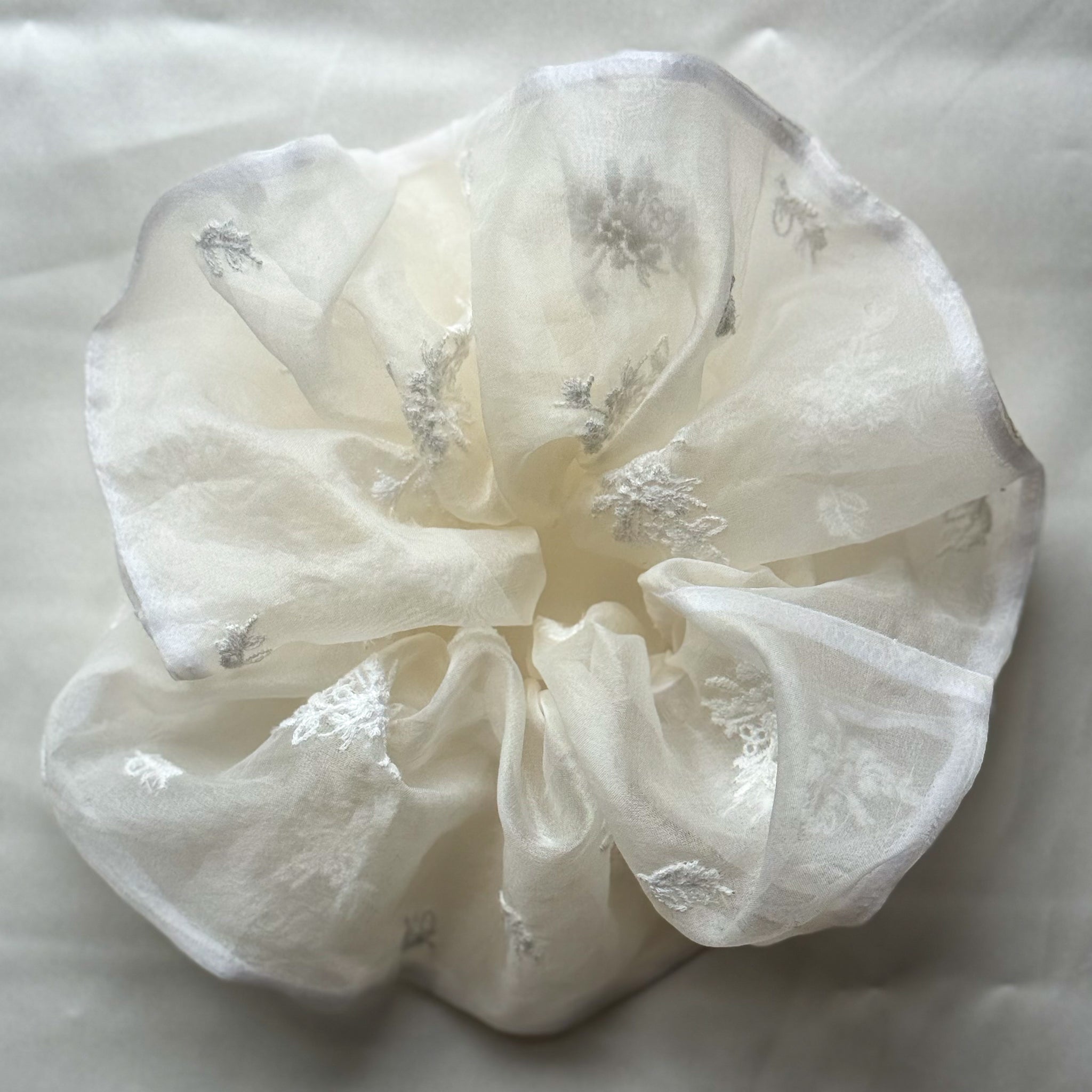 An off-white sheer organza hair scrunchie with delicate white floral embroidery, displayed on a soft white fabric background.