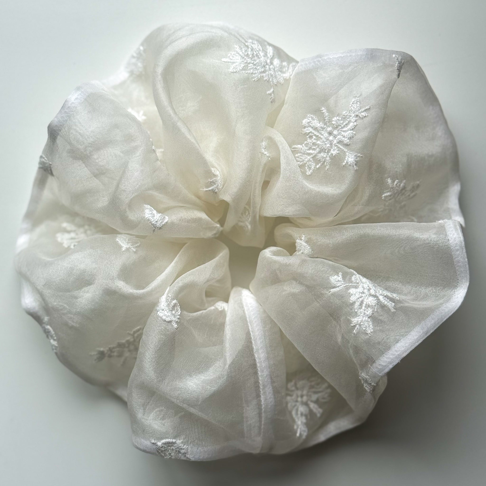 An off-white sheer silk organza hair scrunchie with embroidered flower patterns, photographed on a neutral light gray background.