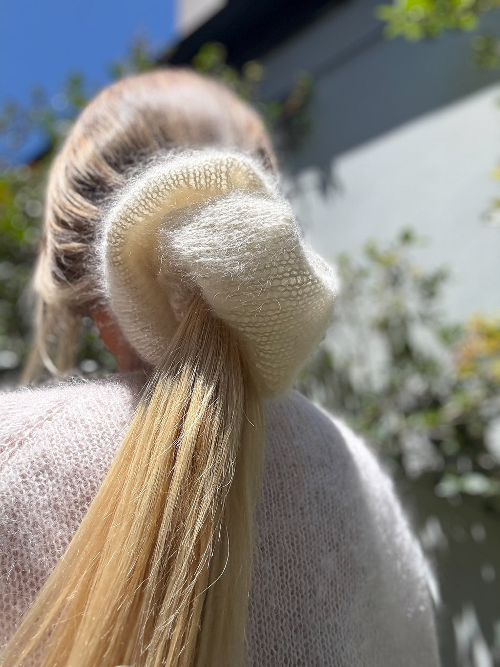 Close-up of a person with blonde hair tied in a ponytail with a cream scrunchie, outdoors.