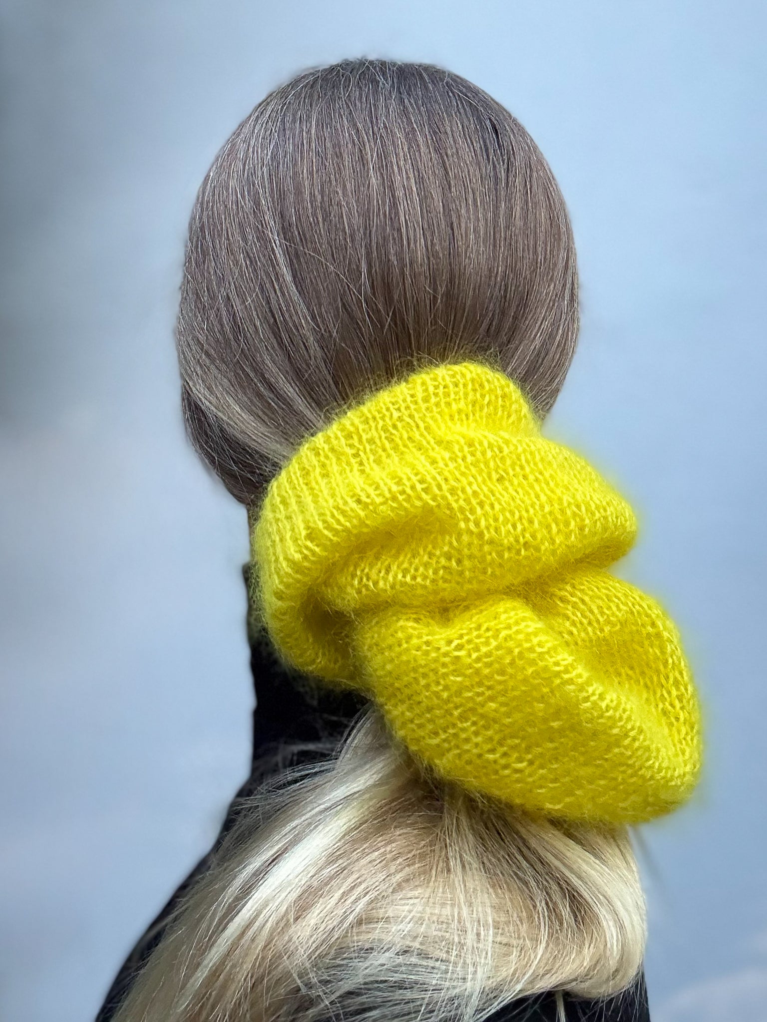 A bright yellow chunky knit scrunchie holding up a blonde and brown ponytail, shown from the back on a model against a soft gray background.