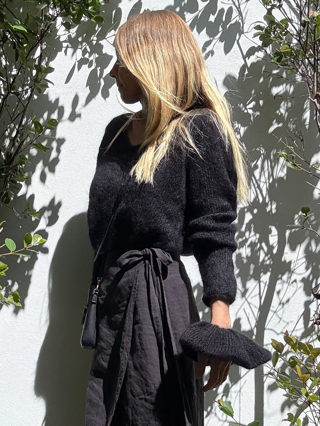 A woman with blonde hair stands in profile against a white wall with leaf shadows, wearing a dark fuzzy knit sweater and a dark linen wrap skirt, wearing a matching massive black knitted scunchie ommher wrist.