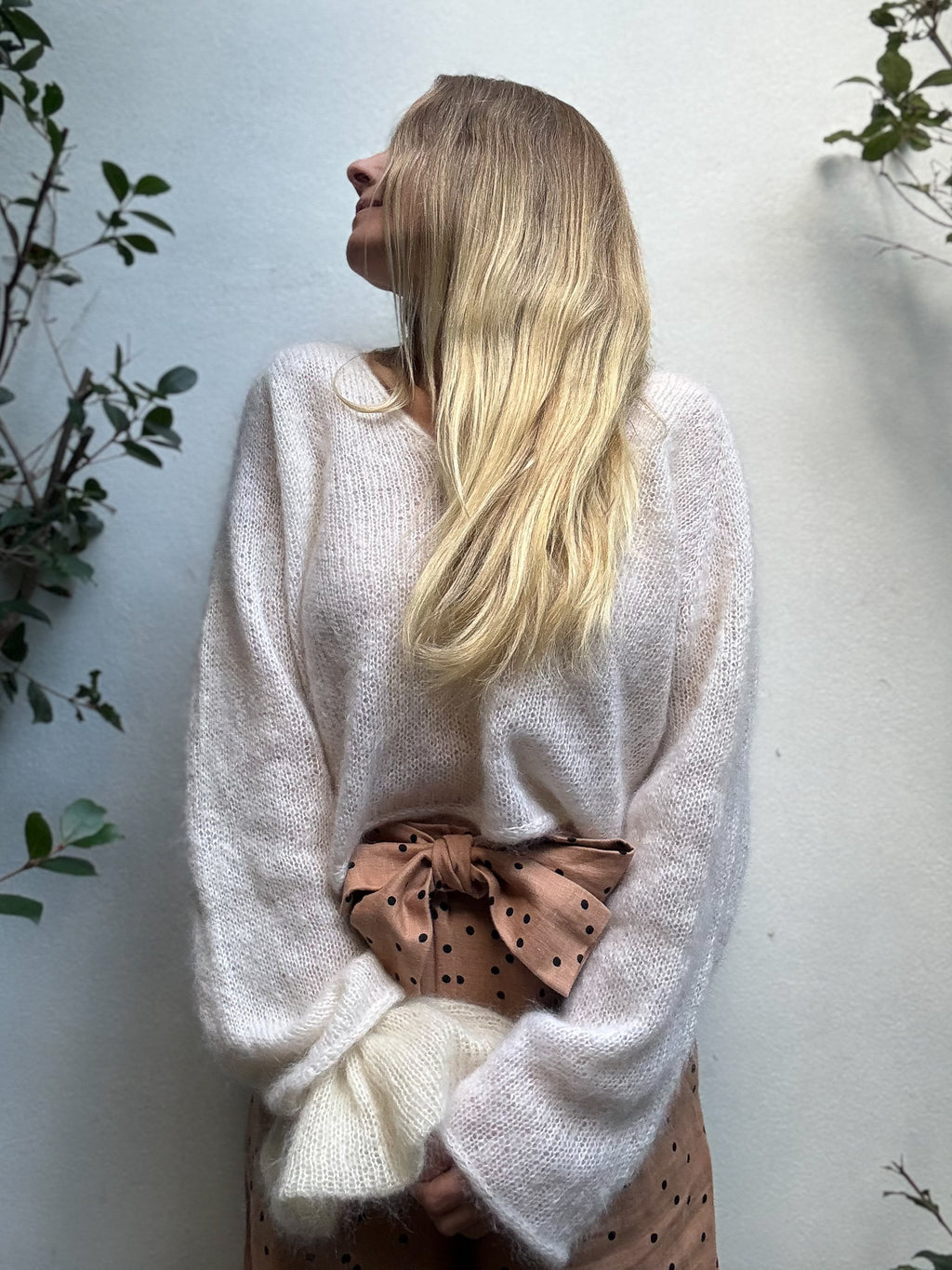 A woman with long blonde hair is wearing a fuzzy off-white long-sleeve sweater and a brown polka-dot pants with a matching bow belt, standing against a light gray wall with green plants and wears an extra large  hand-knitted scrunchie on her wrist.