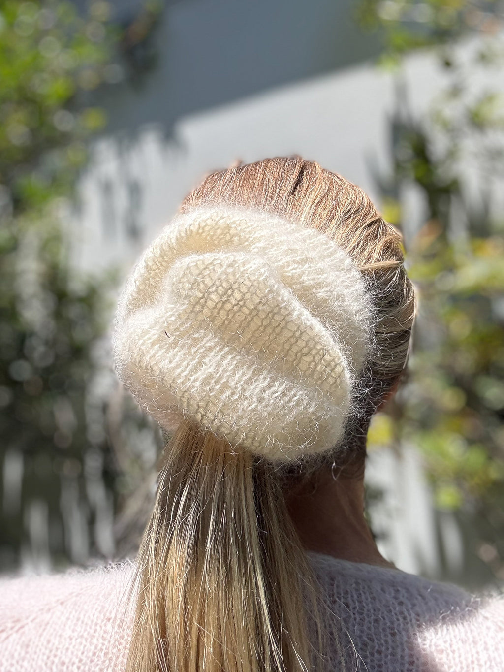 Person wearing a cream-colored knit scrunchie with blurred greenery in the background