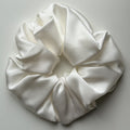 A large white silk satin hair scrunchie with a soft, lustrous sheen, photographed from a top-down angle on a light gray background.