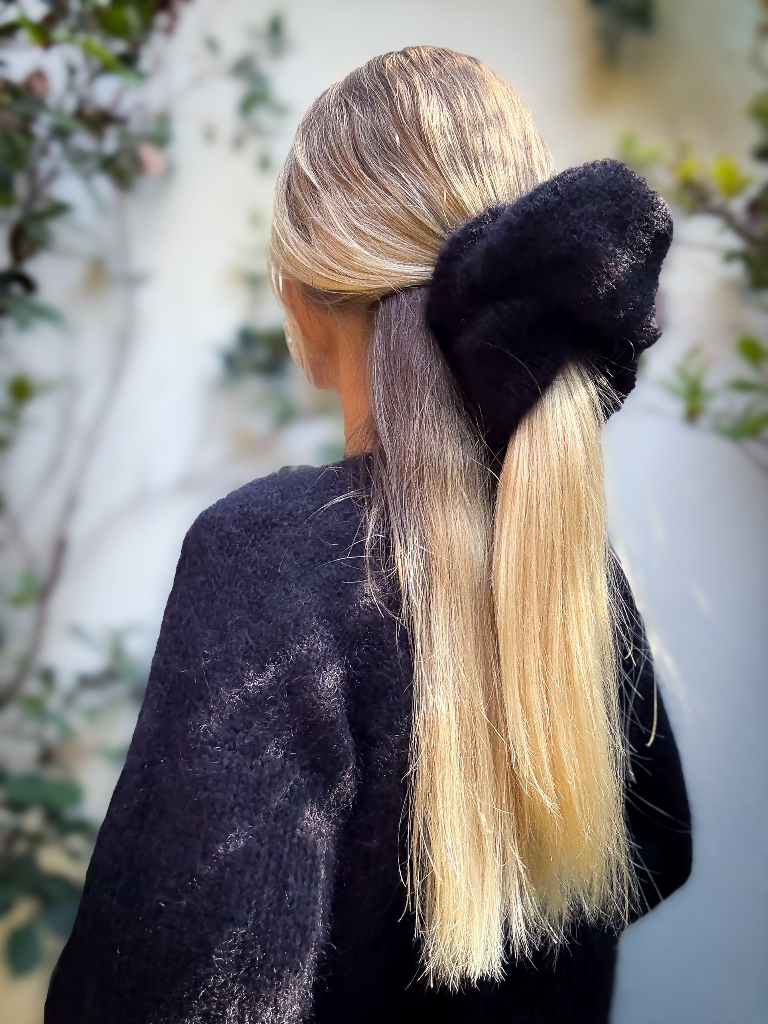 Person with blonde hair tied in a ponytail with a black hand-knitted extra large hair scrunchie, wearing a dark jumper outdoors.