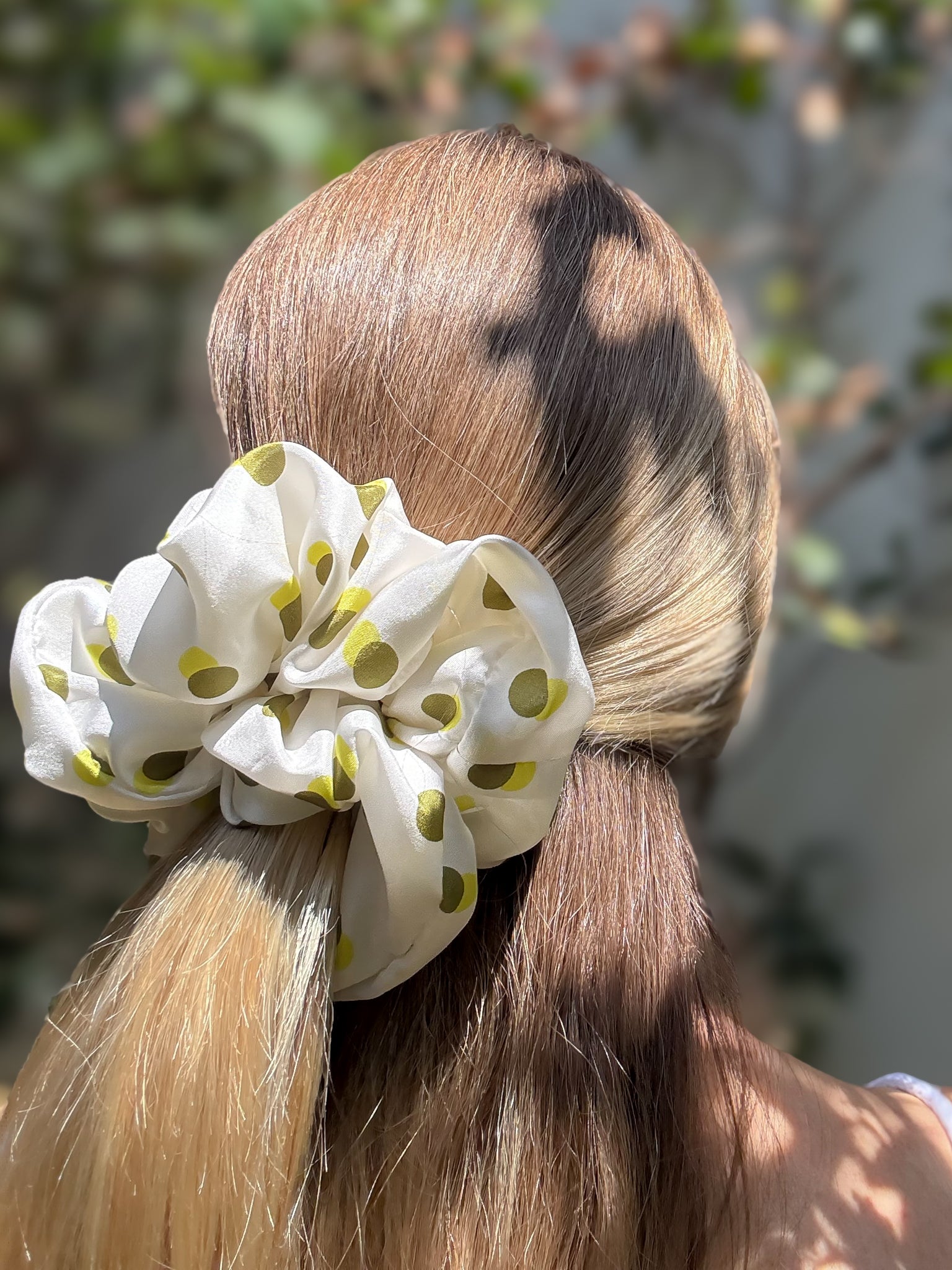 White pure silk scrunchie with green polka dots on a ponytail against a blurred natural background