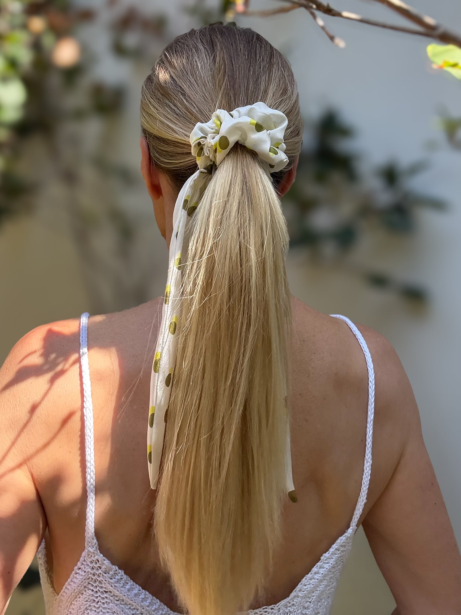 Woman with a high ponytail wearing a scrunchie, standing outdoors with greenery in the background.