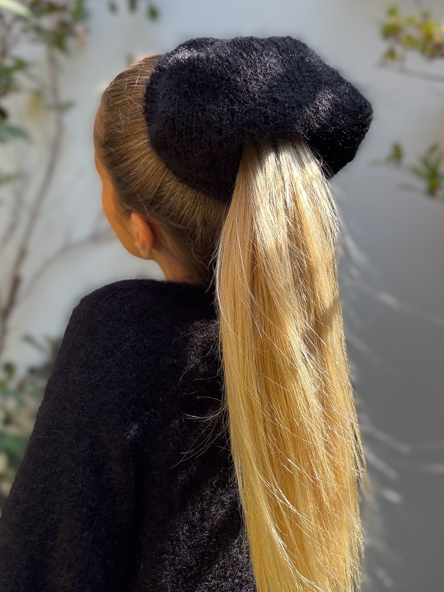 Person with a black extra large hand-knitted hair scrunchie holding a ponytail against a blurred outdoor background