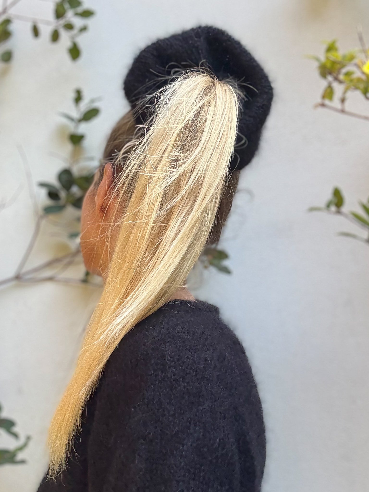 Person with long blonde hair styled in a ponytail with a giant black hair scrunchie wearing a matching black jumper against a light background.