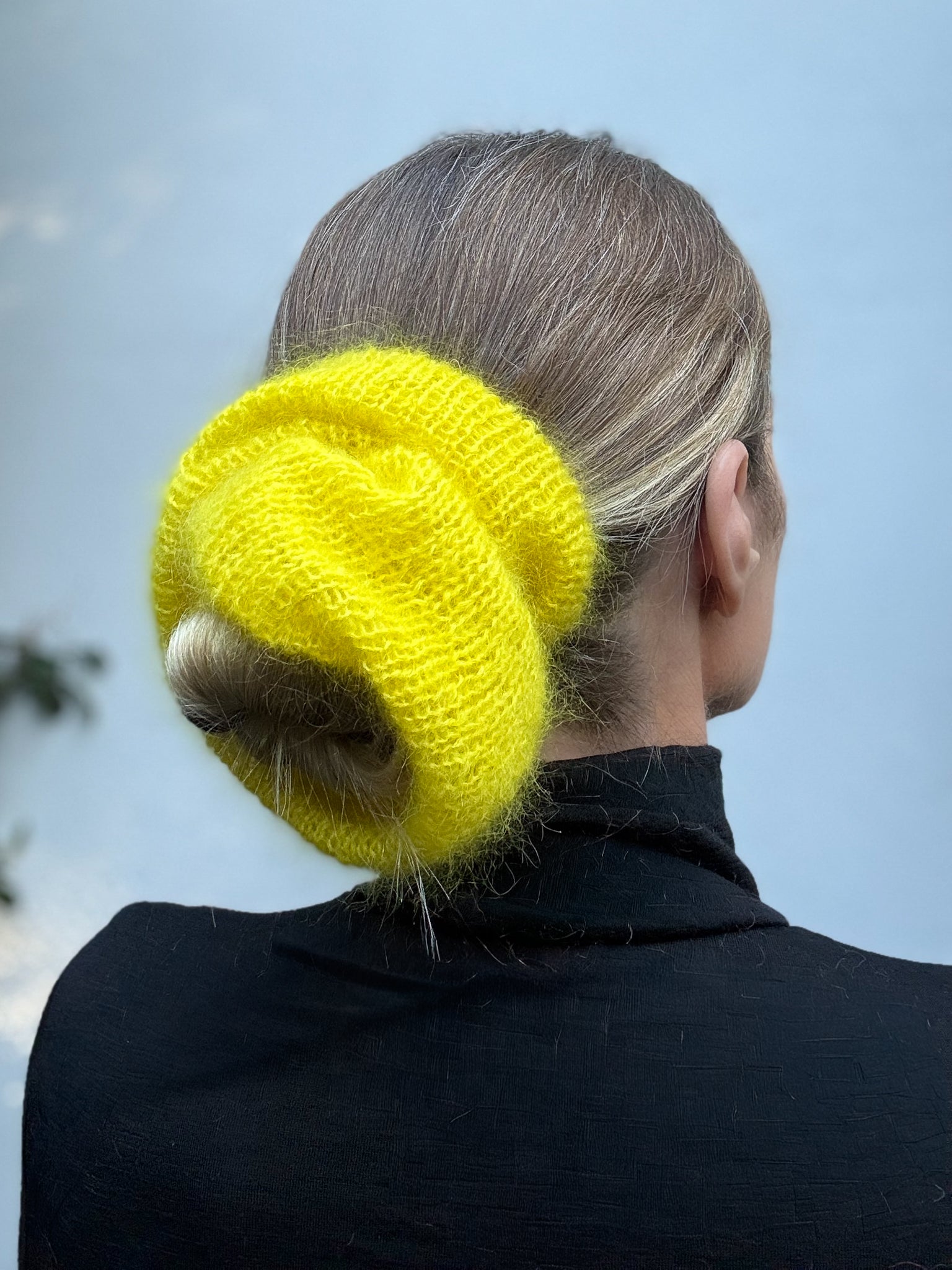 A large bright yellow chunky knit hair scrunchie worn by a woman seen from behind, with her hair in a low bun and a black top against a pale blue sky background.