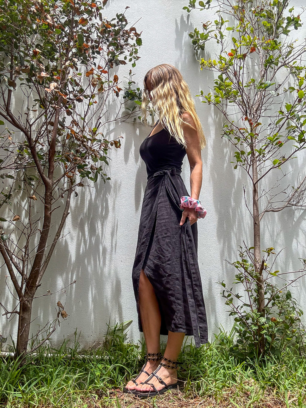 A woman stands outdoors against a white wall with small trees and grass, wearing a sleeveless black midi dress with a side slit and tied waist, paired with black strappy sandals and a large silk scrunchie on her wrist.