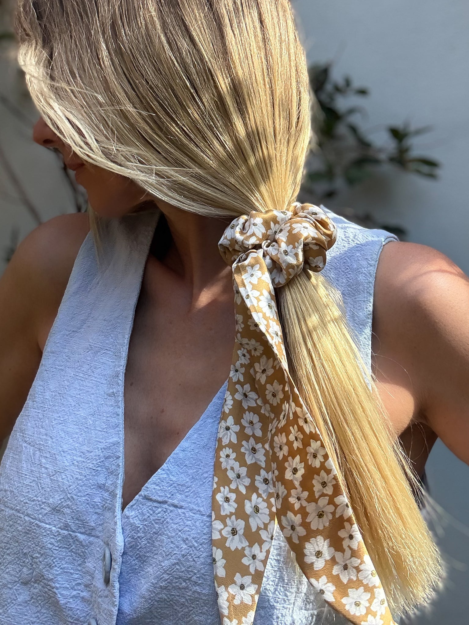 Person with blonde hair tied with a floral silk scrunchie against a blurred outdoor background