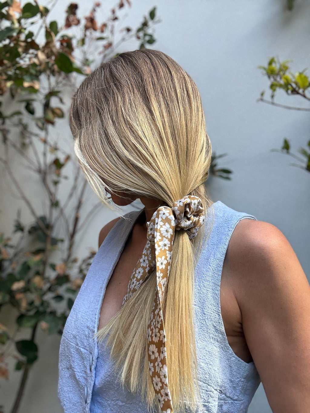Person with blonde hair tied in a low ponytail with a classic silk scrunchie, wearing a light blue vest.