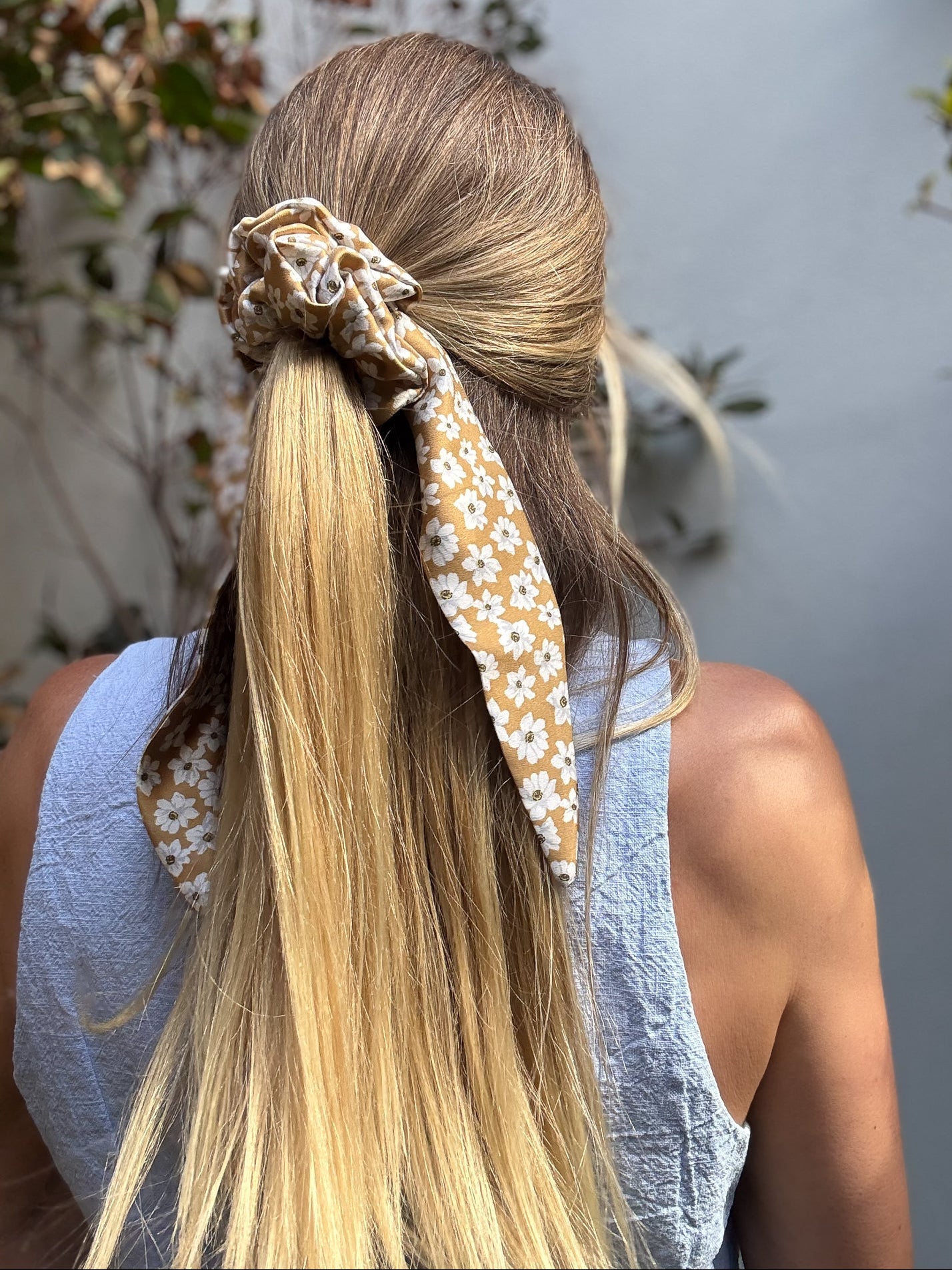 Person with long blonde hair tied back with a floral silk scrunchie, wearing a light blue vest.