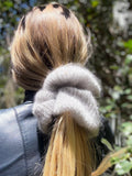 Close-up of a blonde ponytail with a large gray hand knitted scrunchie against a blurred natural background
