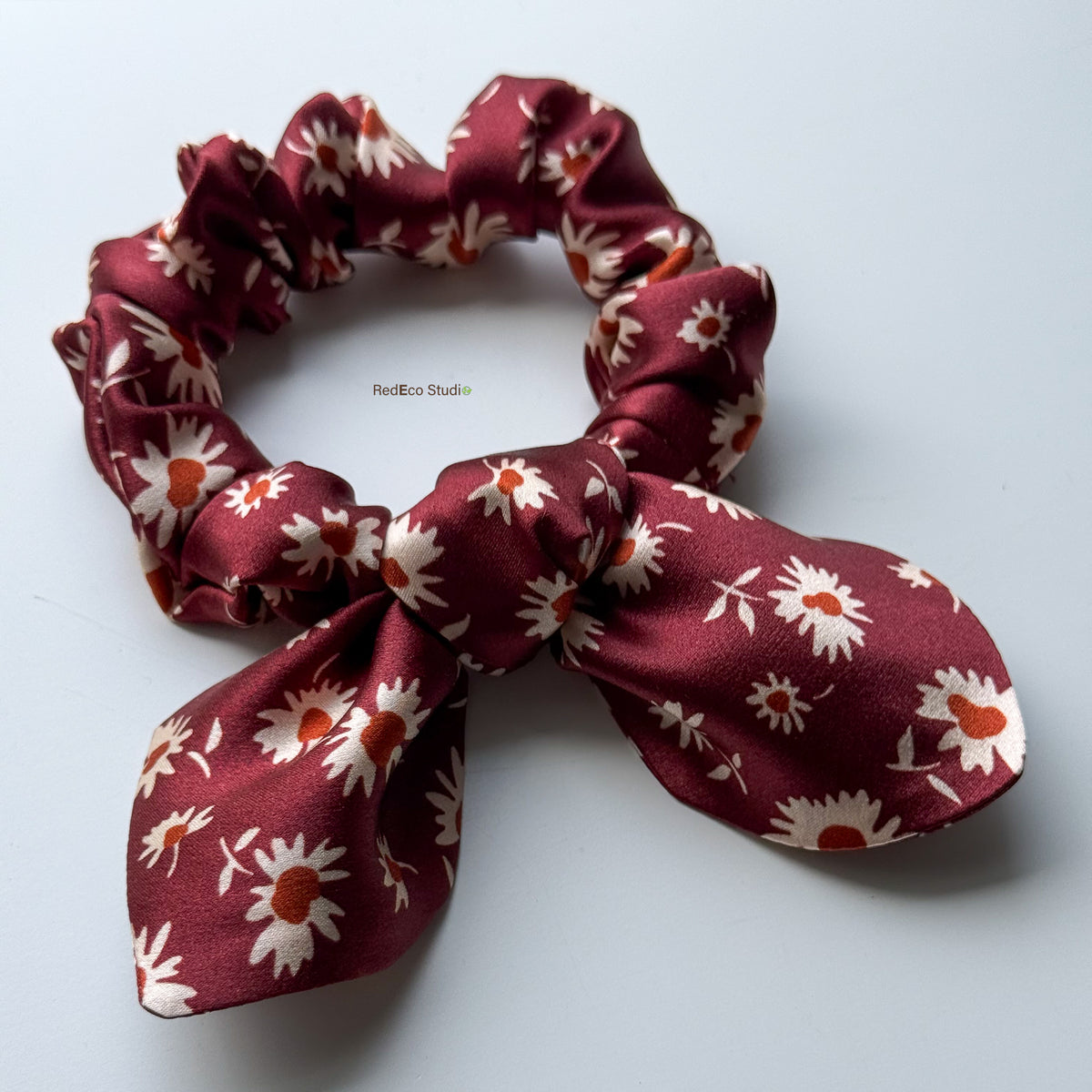 Angled view of the Merlot Daisy mini pure silk scrunchie with its bow tails, showing the satin sheen of the burgundy daisy print