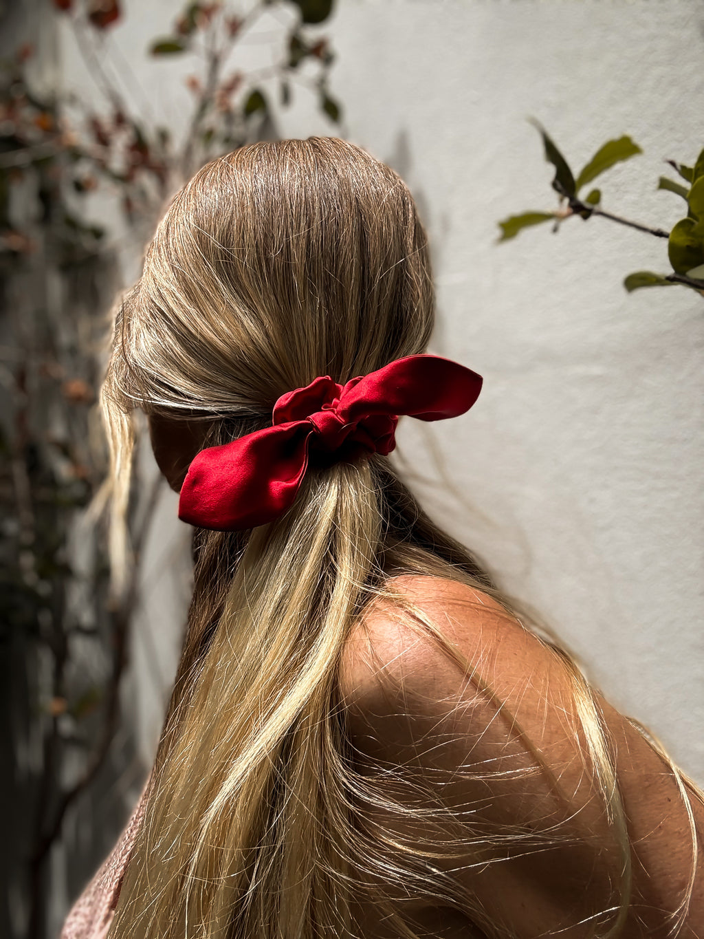 Person with long blonde hair tied back with a red ribbon, standing against a neutral background.