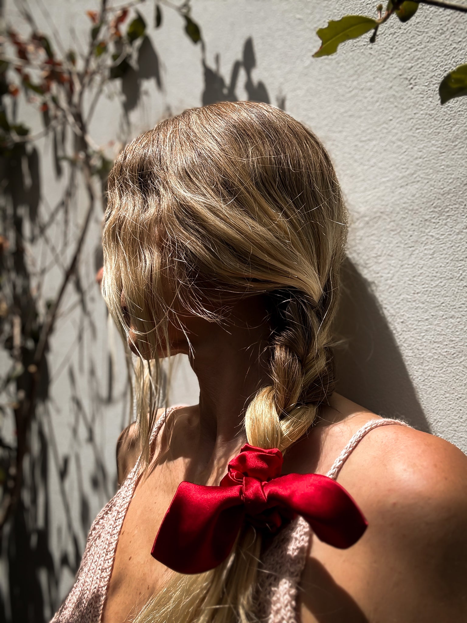 Woman with braided hair and red bow standing against a light-colored wall with tree shadows.