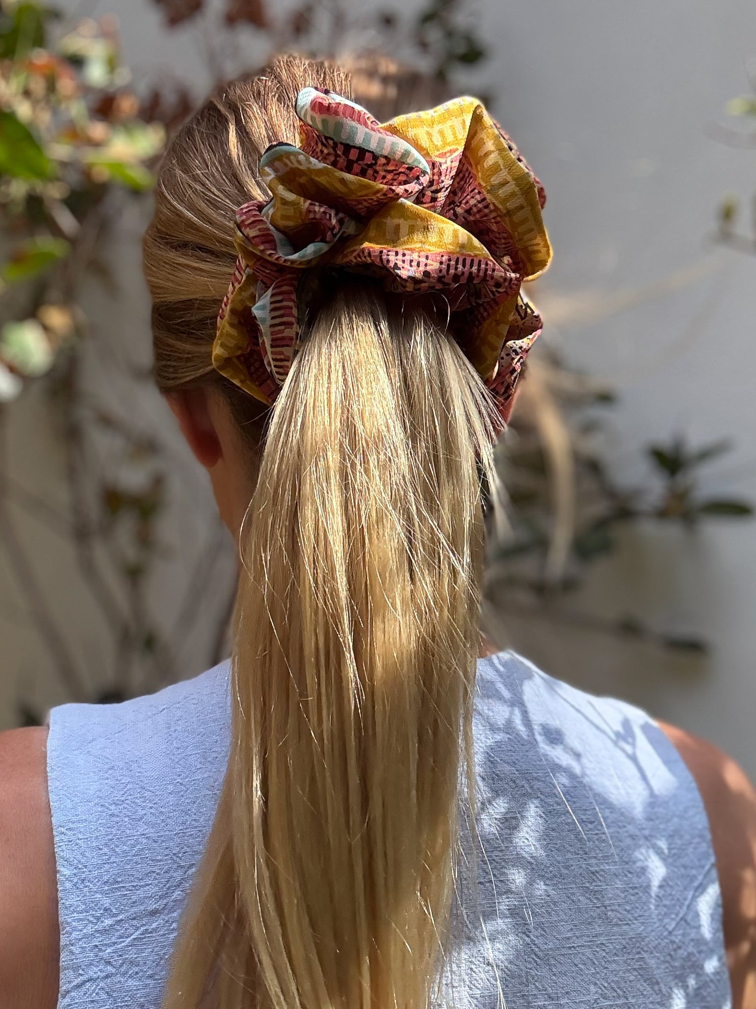 Person with a colorful extra large silk scrunchie in their hair, standing outdoors with plants in the background